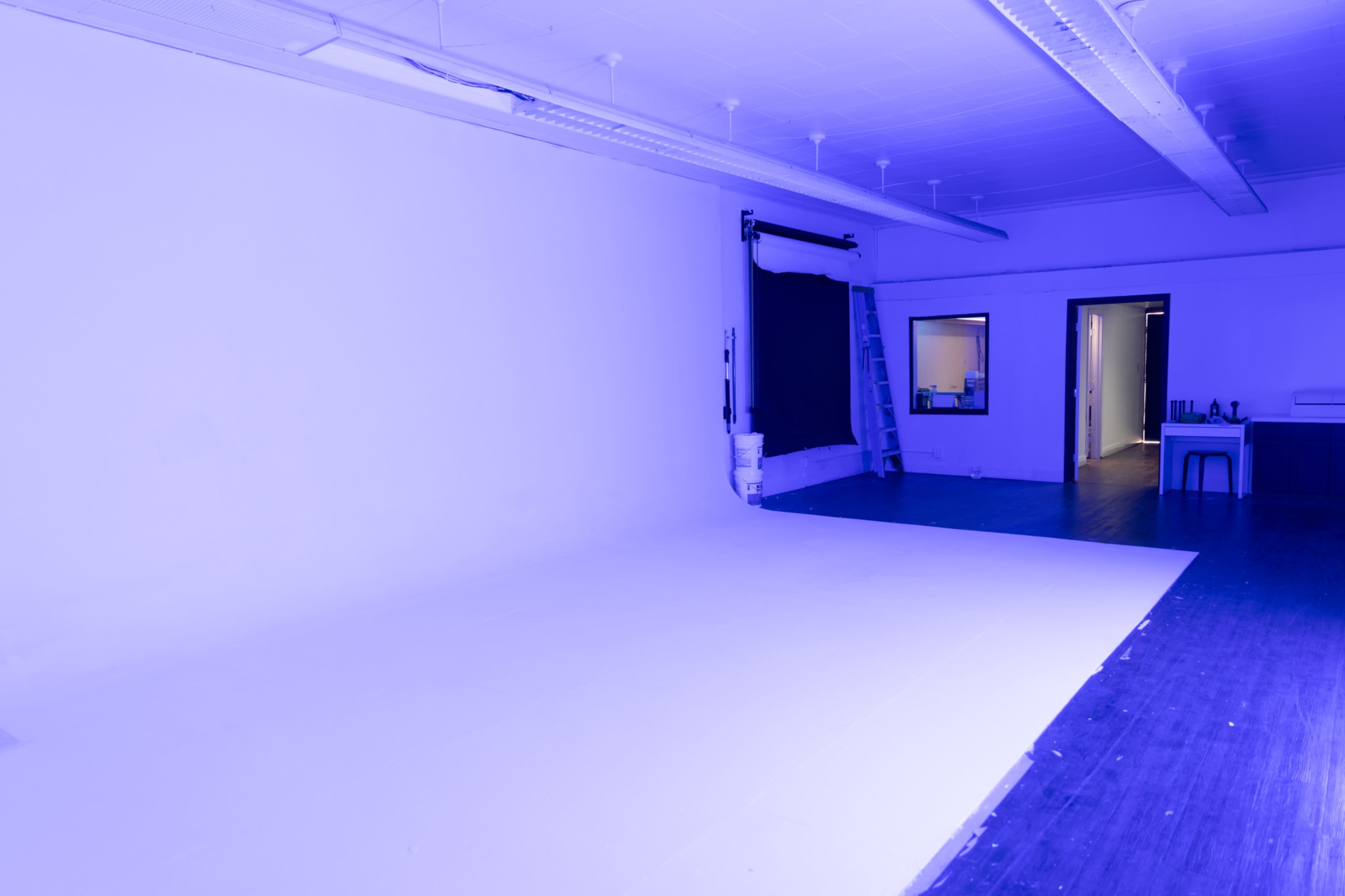 A large, empty room is illuminated in a blue hue, featuring a white backdrop on one wall and a black backdrop hanging from a support on the other side.
