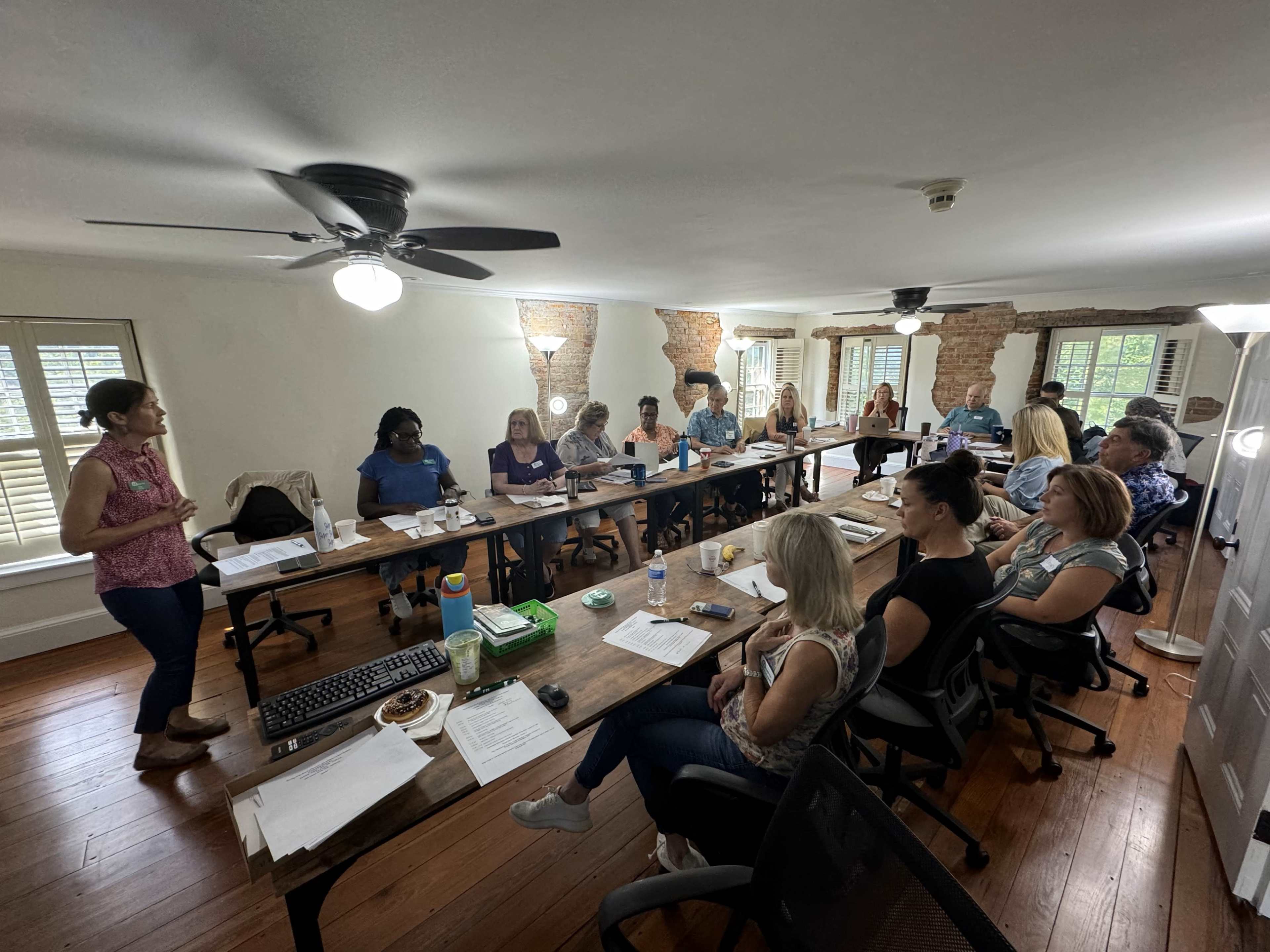 A group of people sit around a long conference table in a well-lit room, attentively listening to a speaker at the front.