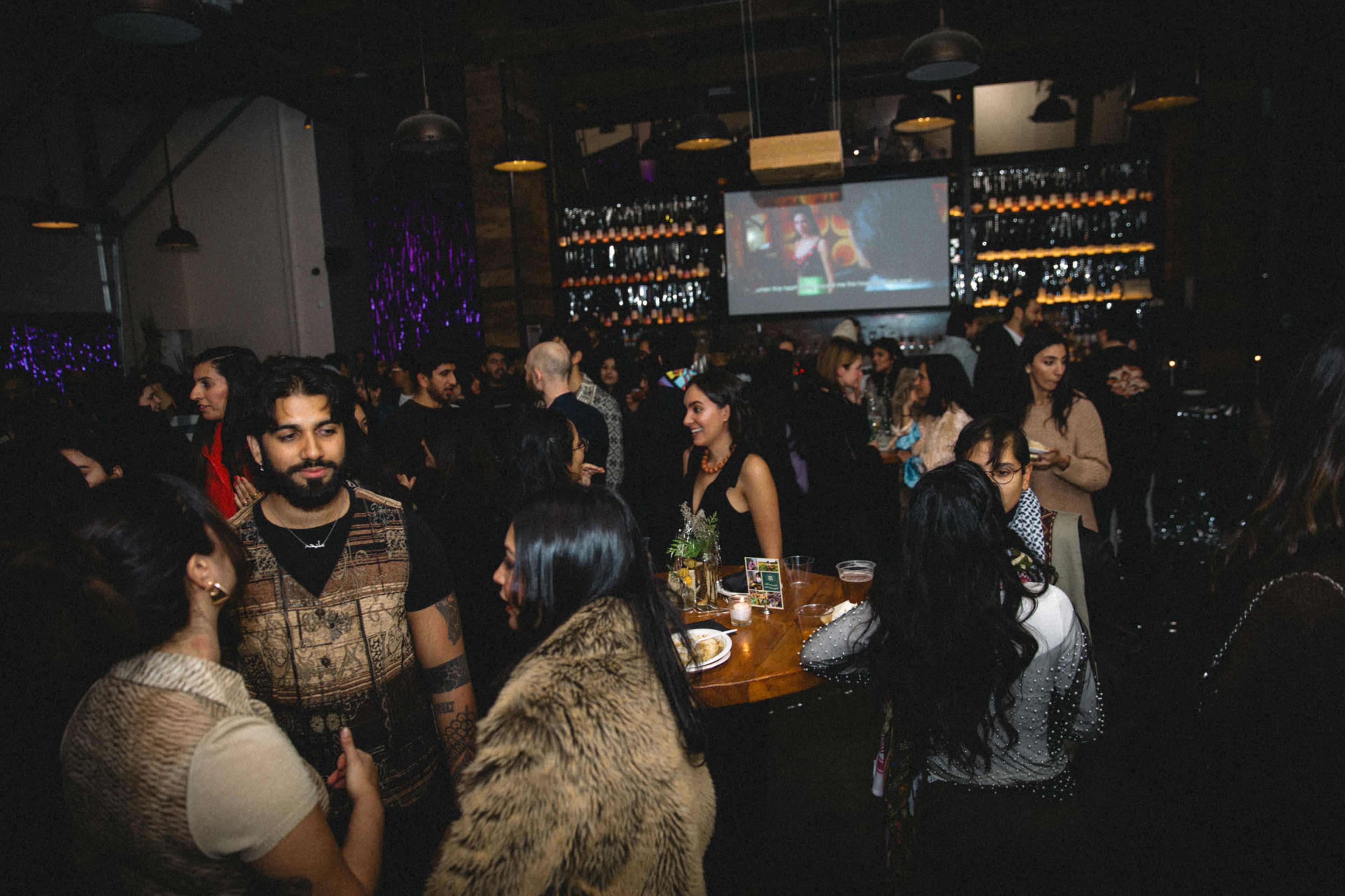 A crowded venue features people socializing and enjoying drinks, with a large screen displaying a video in the background.