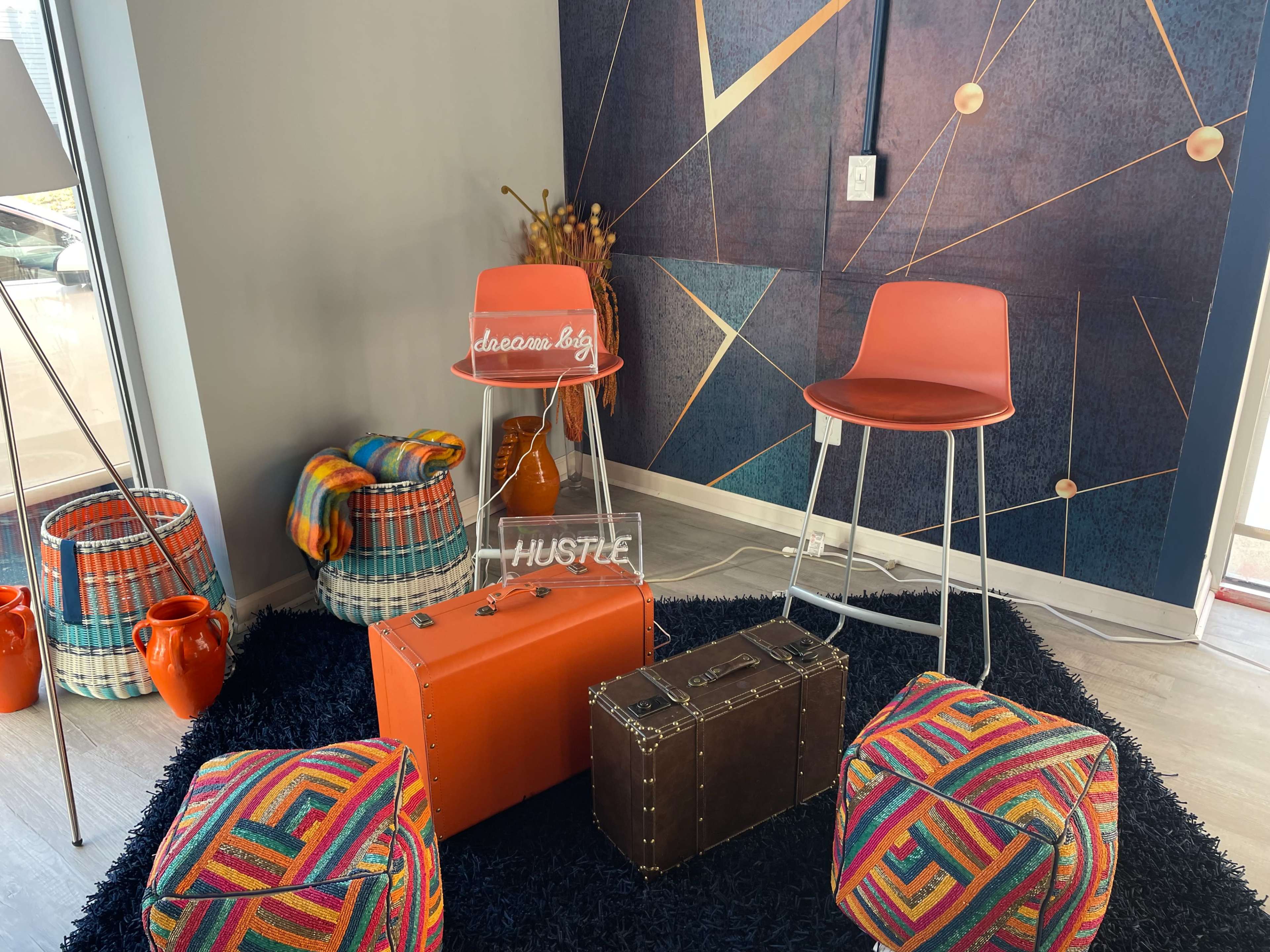 The scene features two orange bar stools, colorful poufs, a vintage-style trunk, and woven baskets against a geometric wall design.