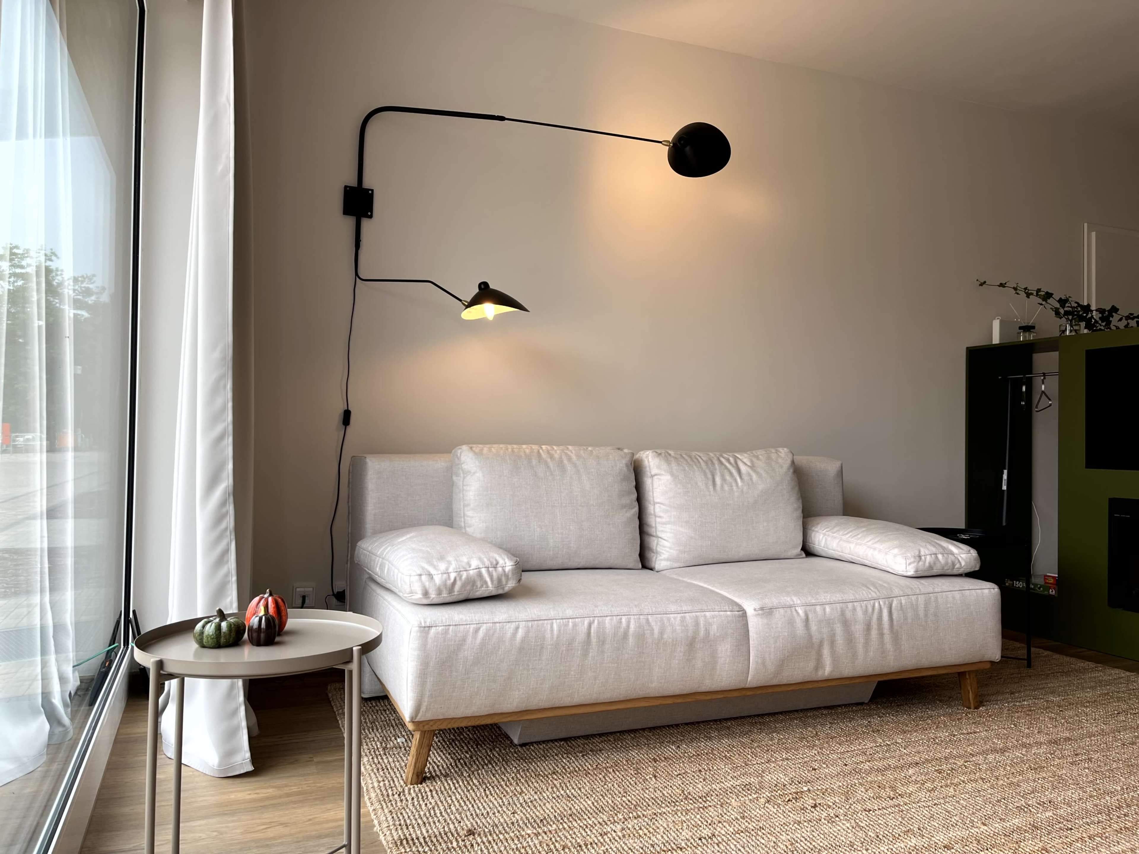 A light gray sofa with two large cushions sits in a brightly lit corner next to a small round table and a wall-mounted lamp.