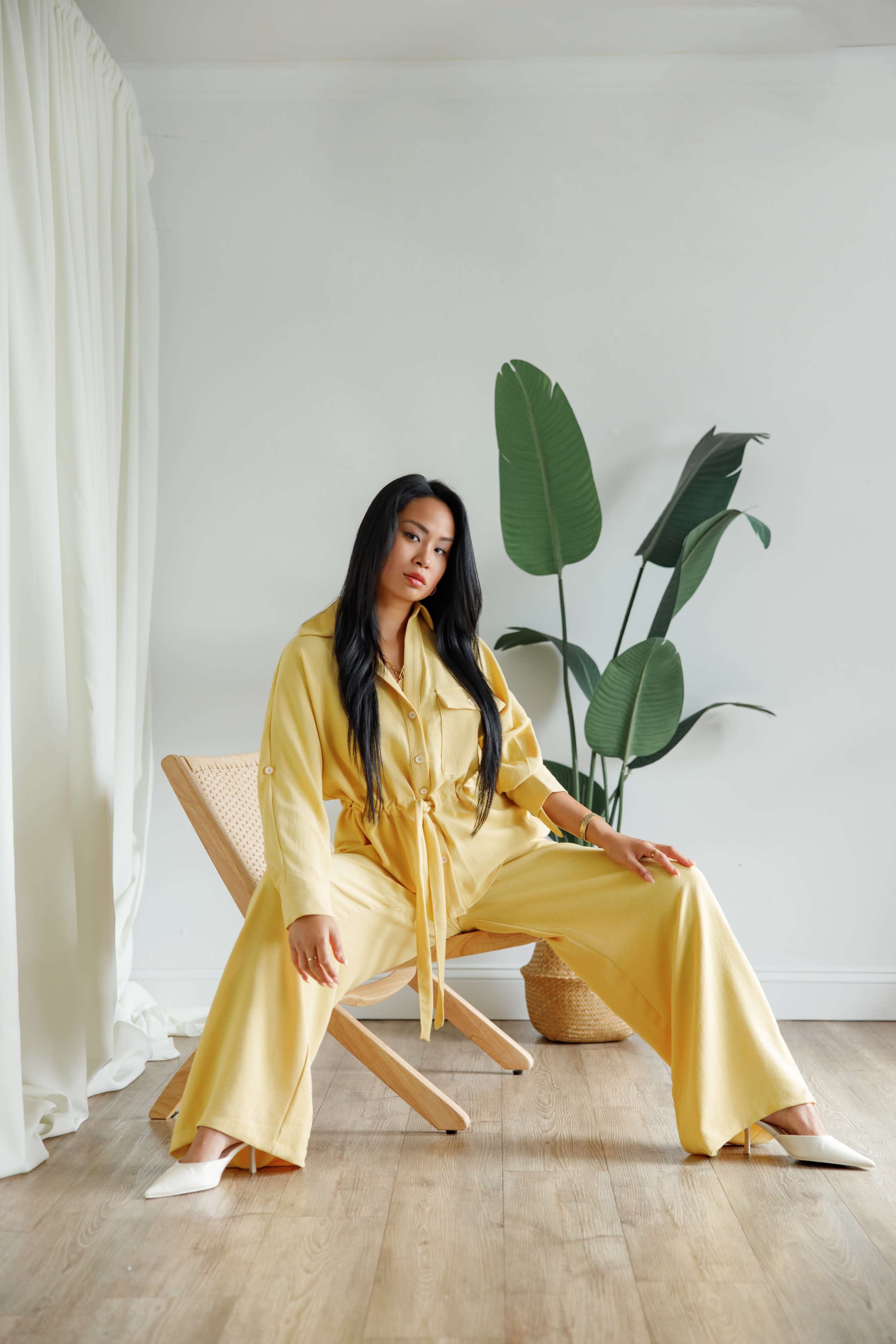 A woman in a yellow outfit sits in a chair with her legs stretched out, beside a large potted plant.