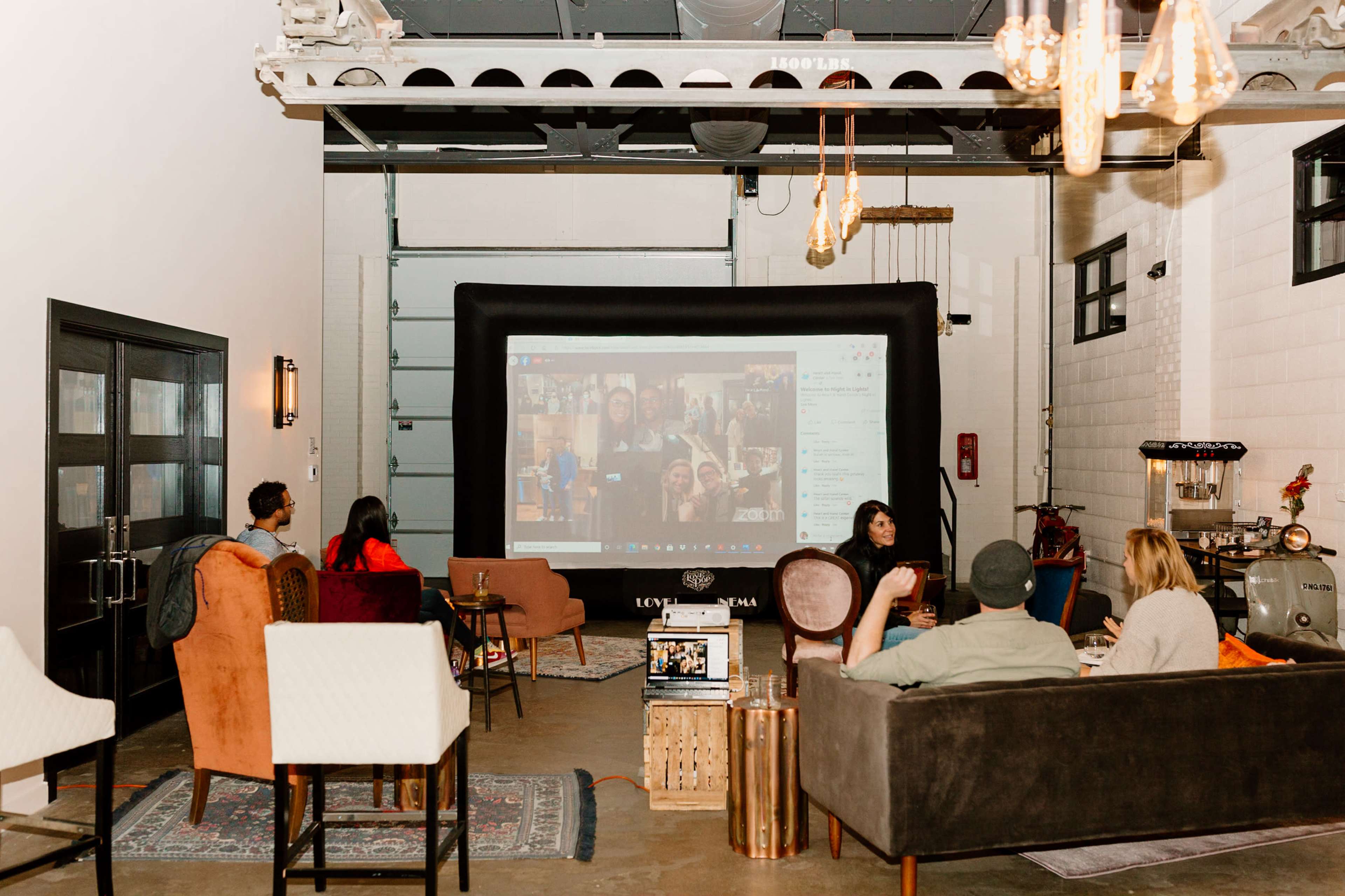 A group of people is seated in a warehouse-style space with a large screen displaying a video call.