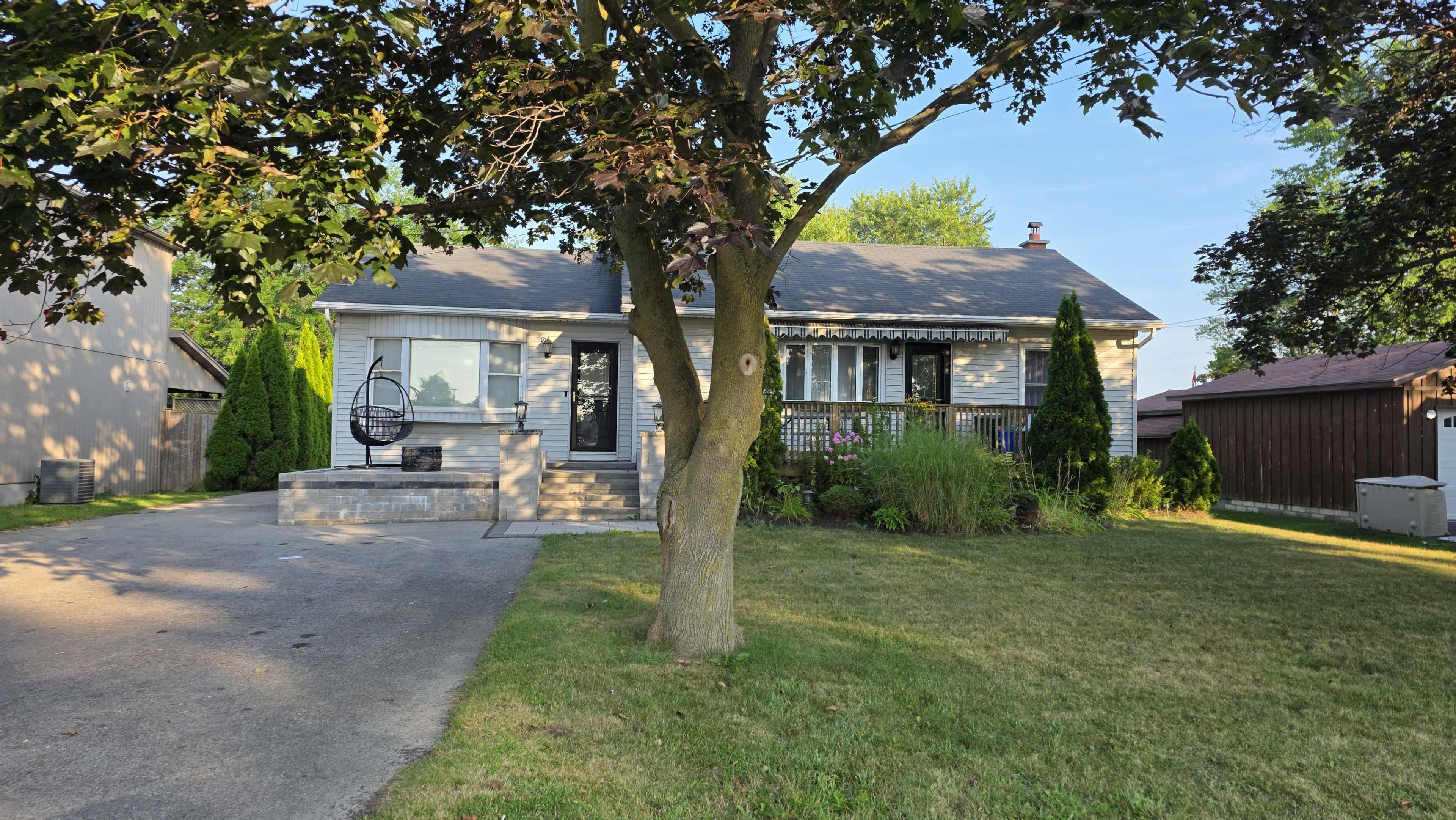 A single-story house with a grey exterior sits on a grassy lot, surrounded by trees and shrubs, with a driveway leading up to the entrance.