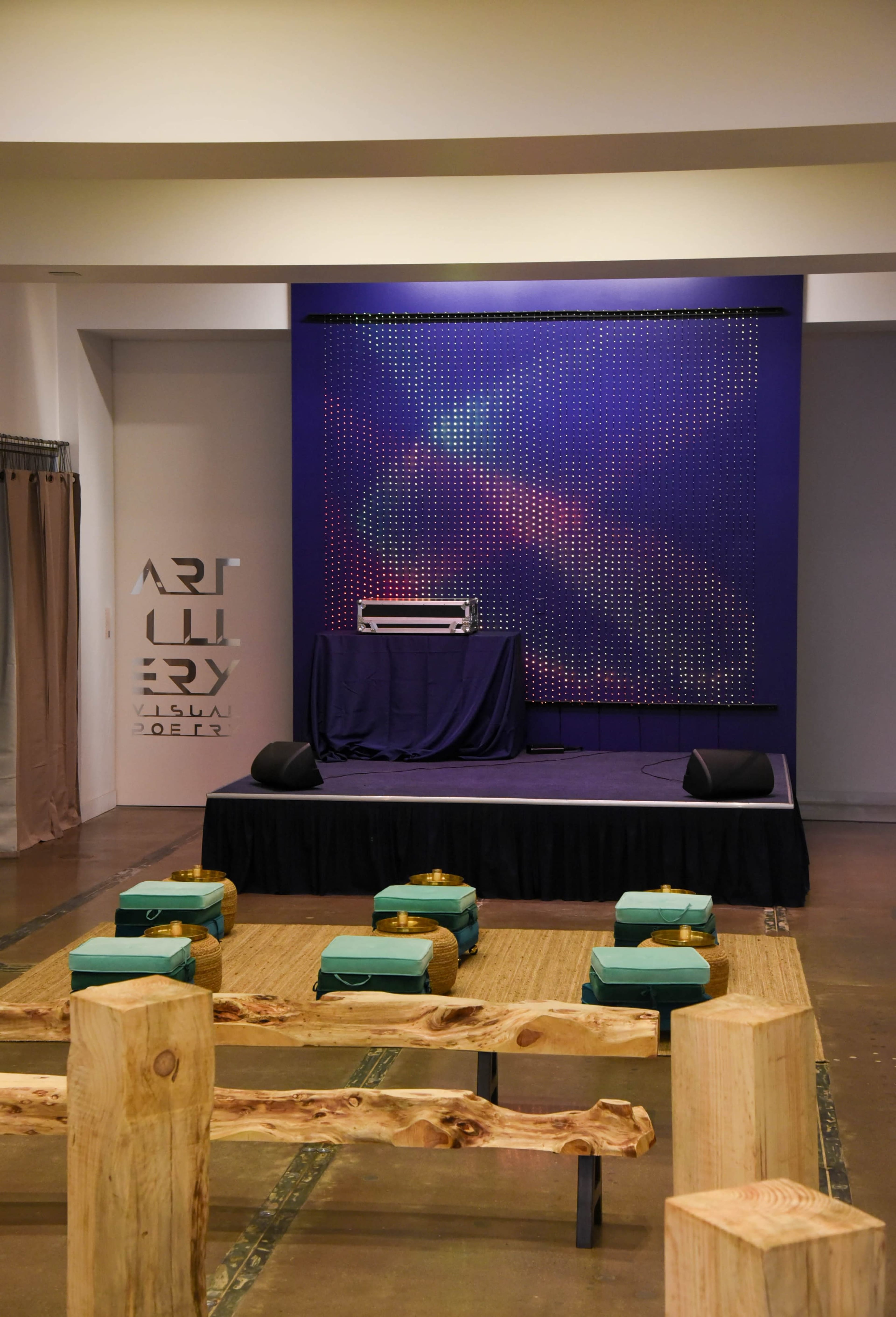 A stage with a patterned backdrop, a table with a sound system, and green seating arranged in front of it on a wooden floor.