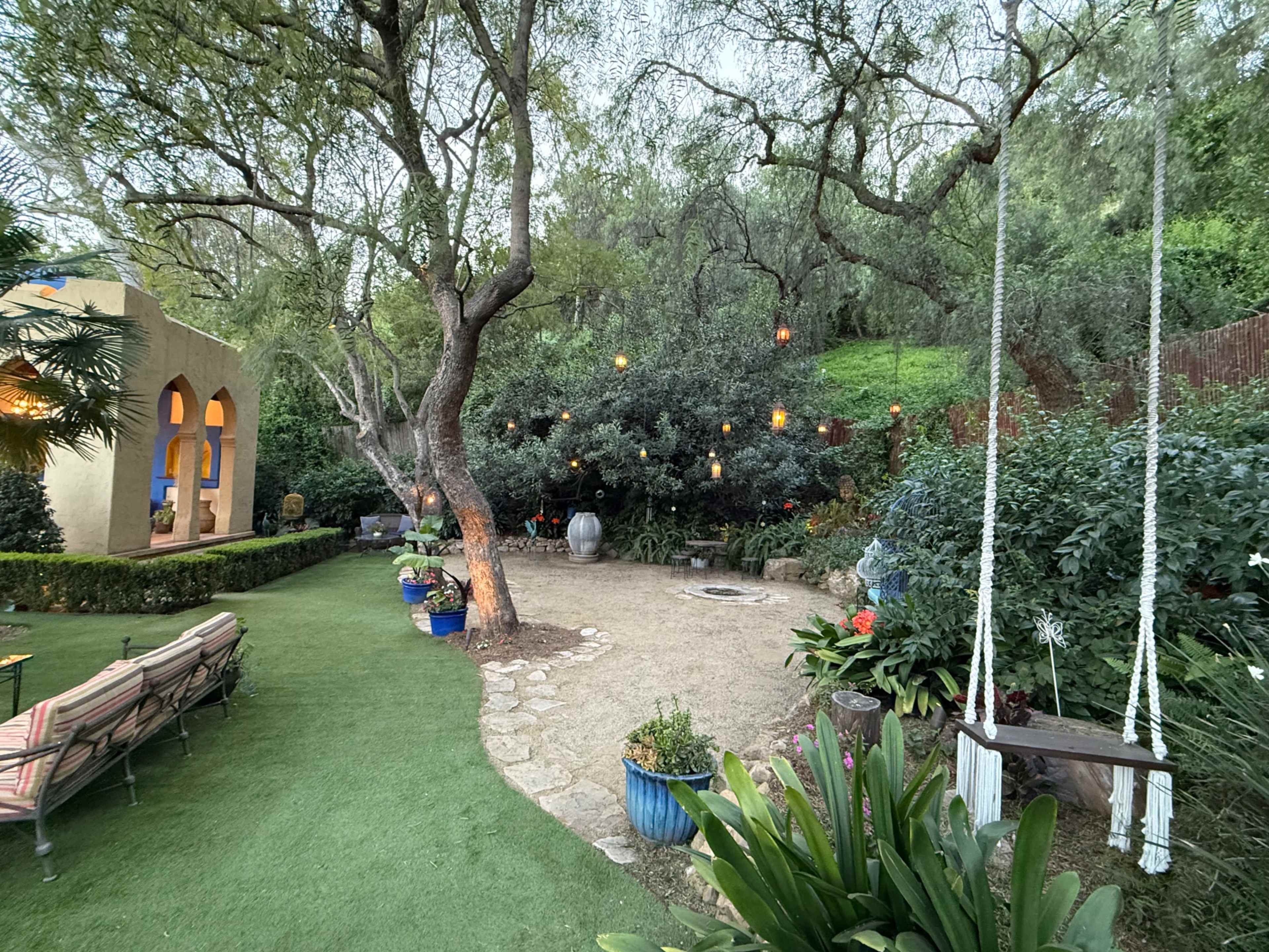 The image shows a tranquil garden with a stone pathway, lush greenery, a swing hanging from a tree, and decorative lighting among the plants.