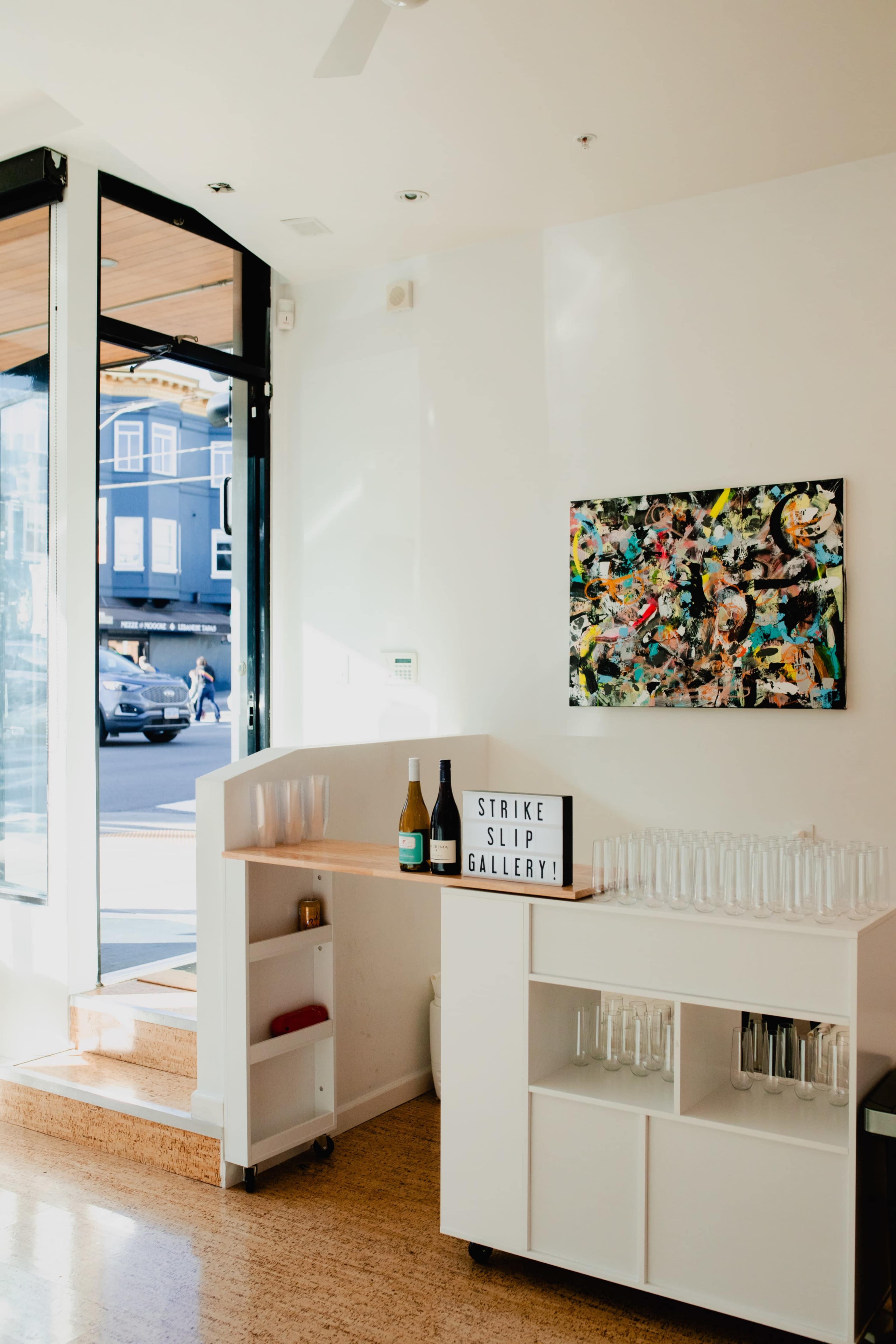 The interior of a bright, modern gallery space featuring a bar area with bottles and glasses, alongside a colorful abstract painting on the wall.