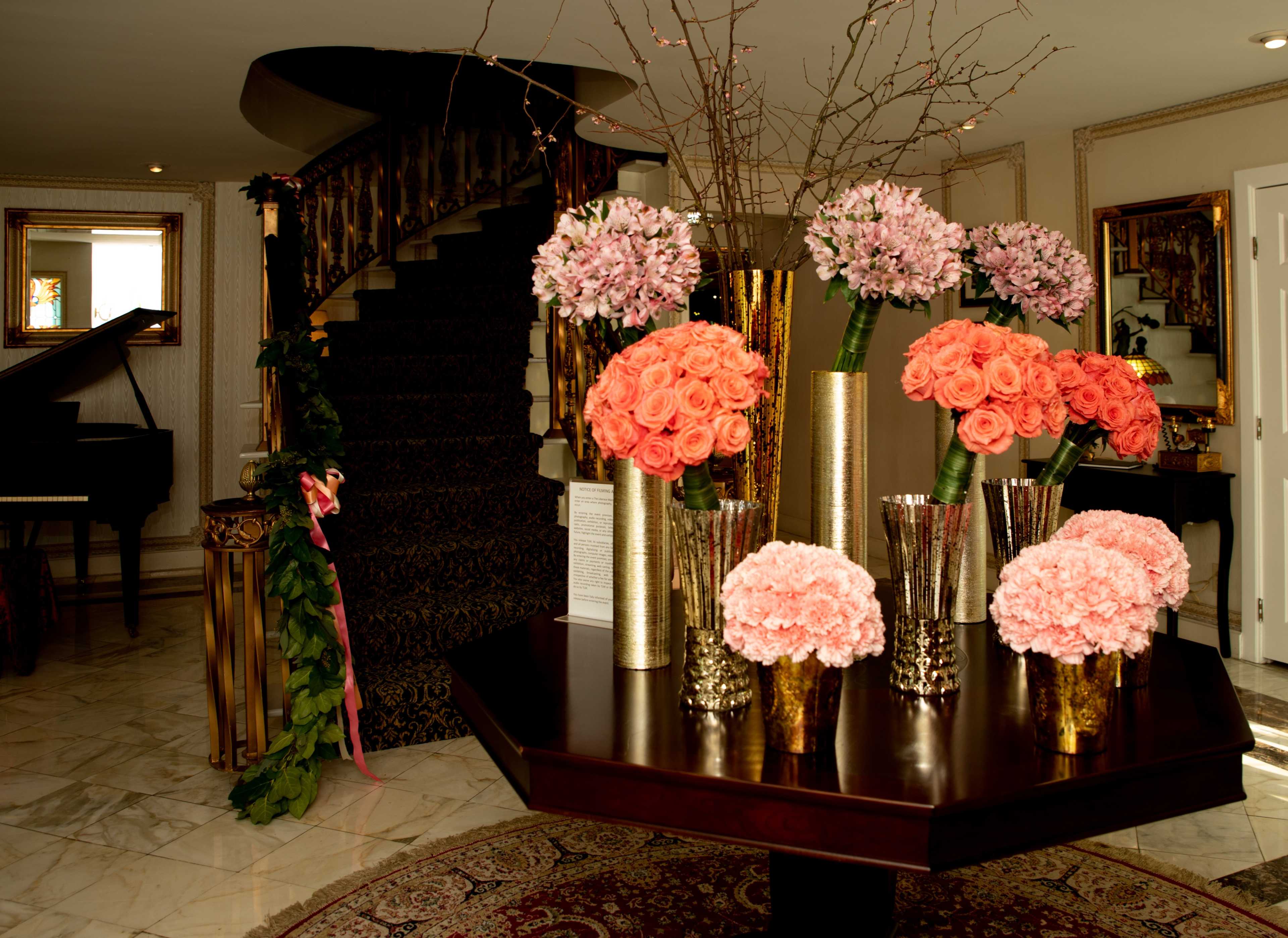 A decorative entryway features a round table with vases of pink and orange flowers, alongside a staircase and a grand piano in the background.