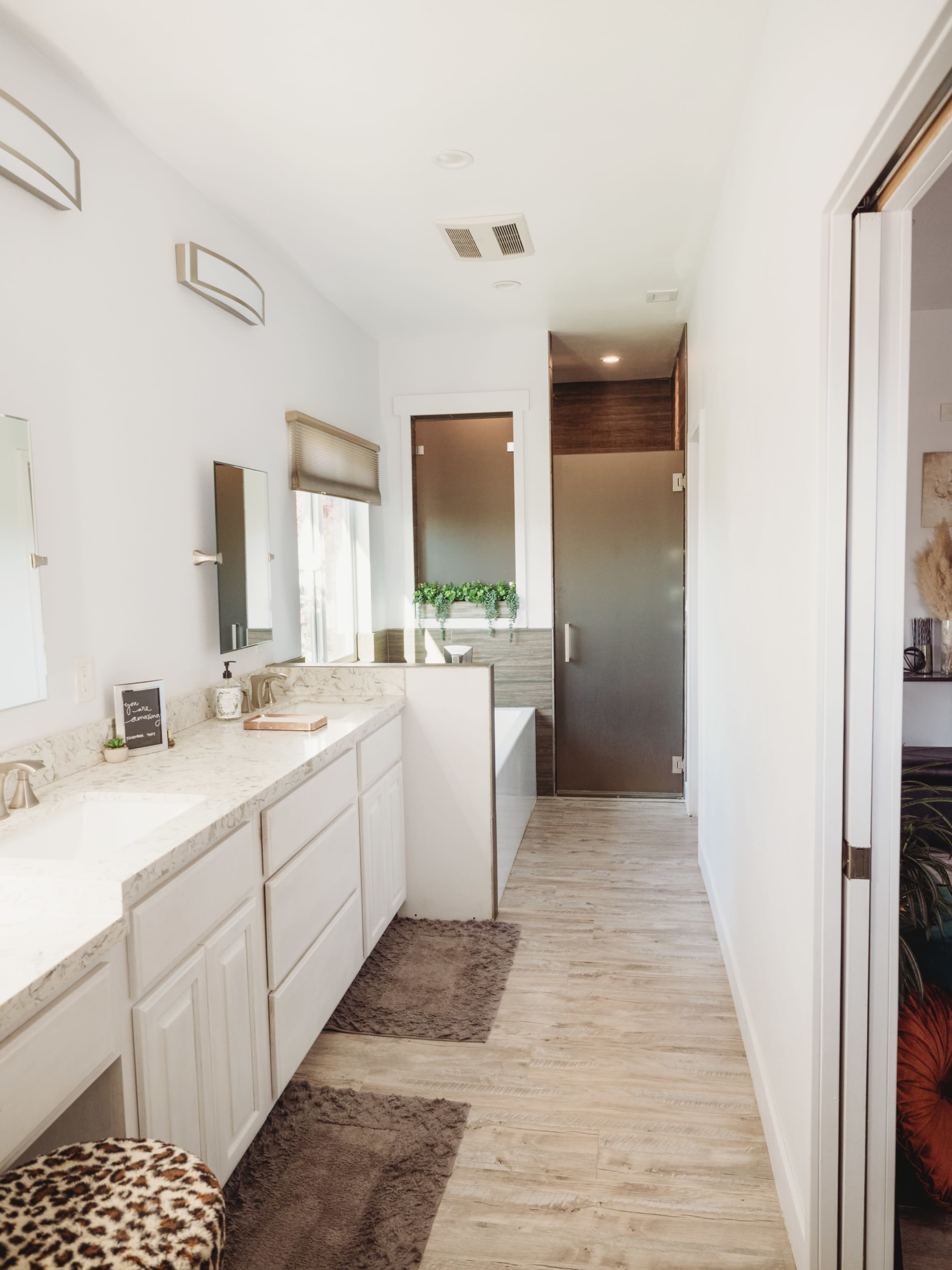A modern bathroom with a double vanity, light-colored cabinets, and a walk-in shower at the end of a hallway.