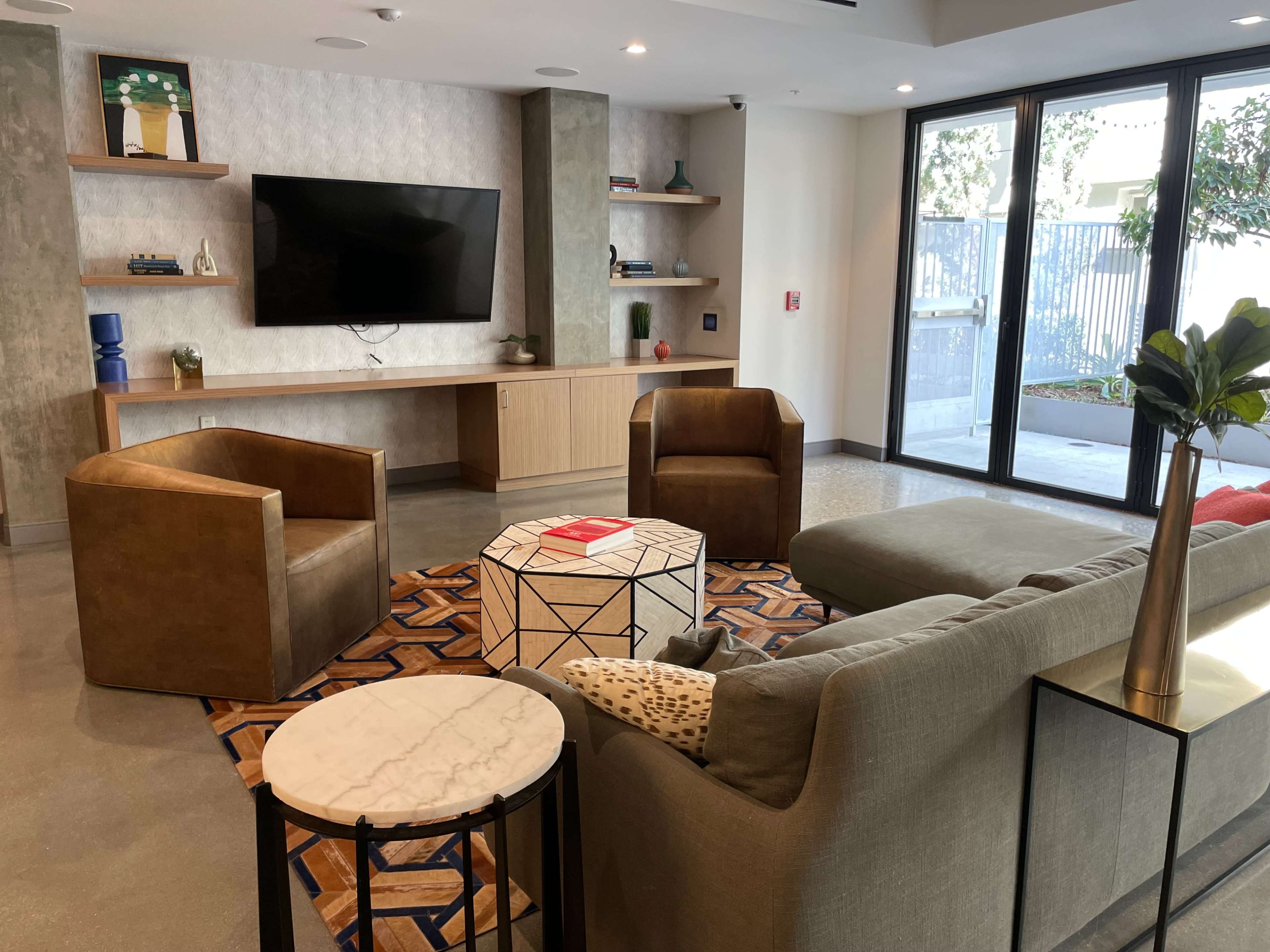 A modern living space featuring two brown chairs, a coffee table, and a large television mounted on the wall, with large windows opening to an outdoor area.