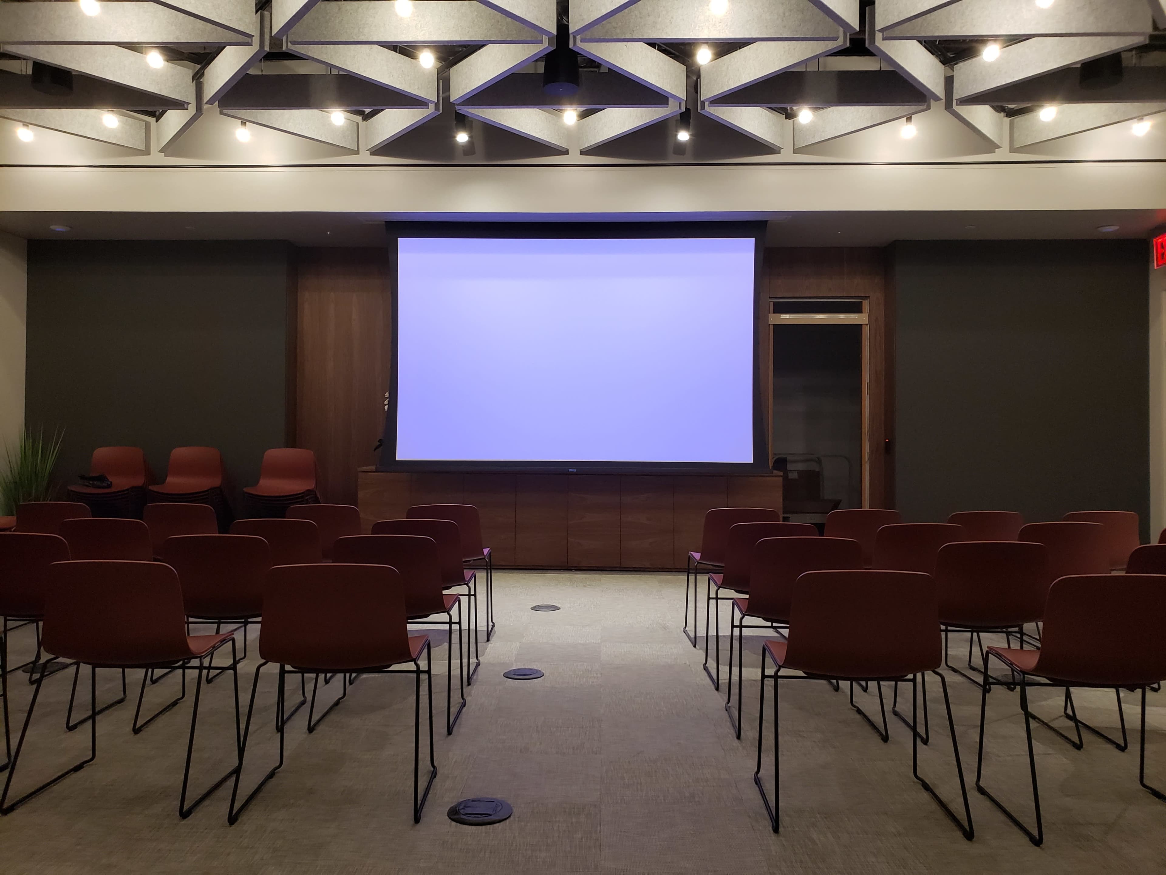A conference room is set with rows of red chairs facing a blank projection screen.