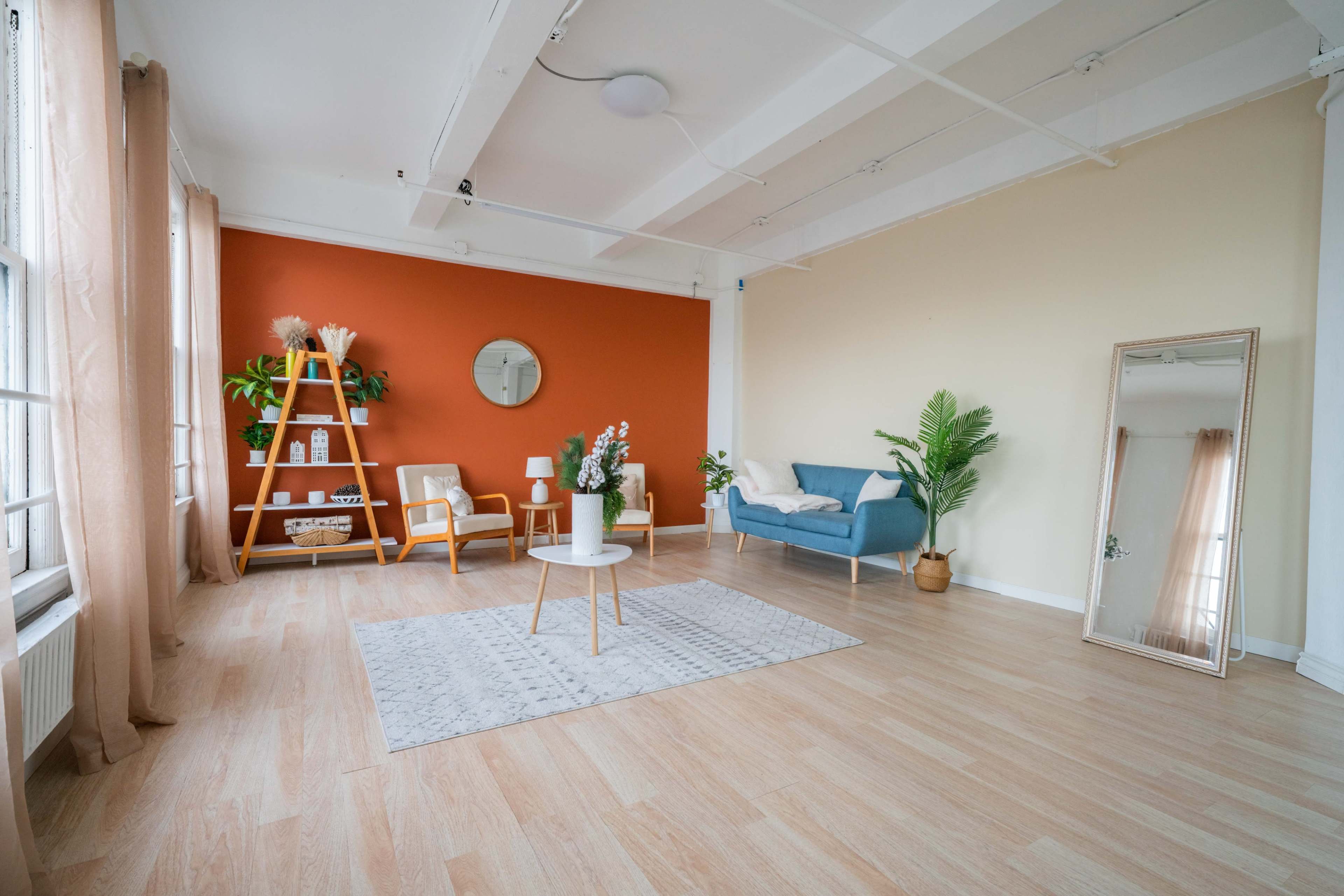 A bright, modern interior living space with an orange accent wall, a blue sofa, wooden shelving, and a large mirror.
