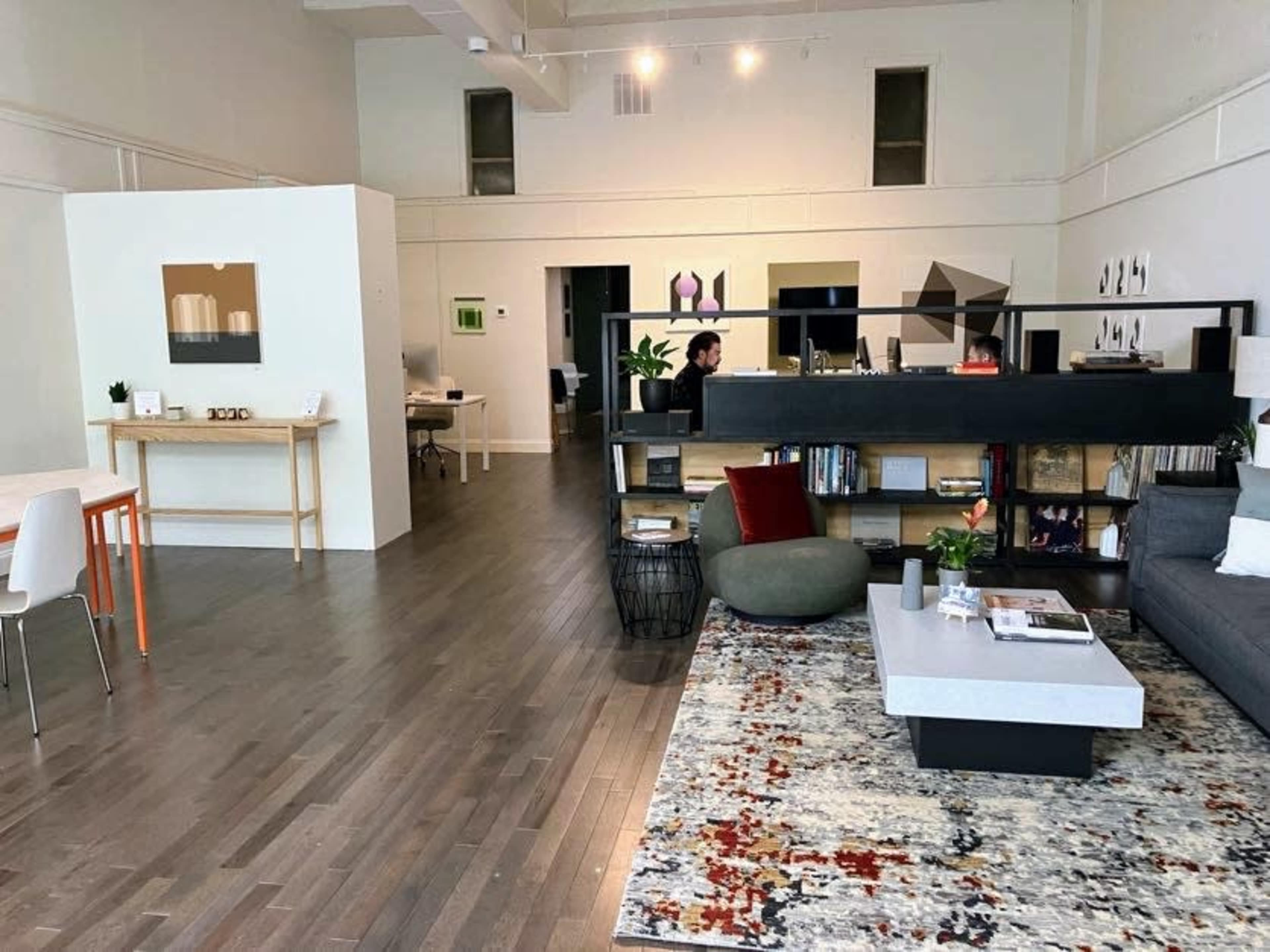 A spacious modern living area featuring a gray sofa, a coffee table on a patterned rug, a bookshelf, and distinct workspaces in the background.