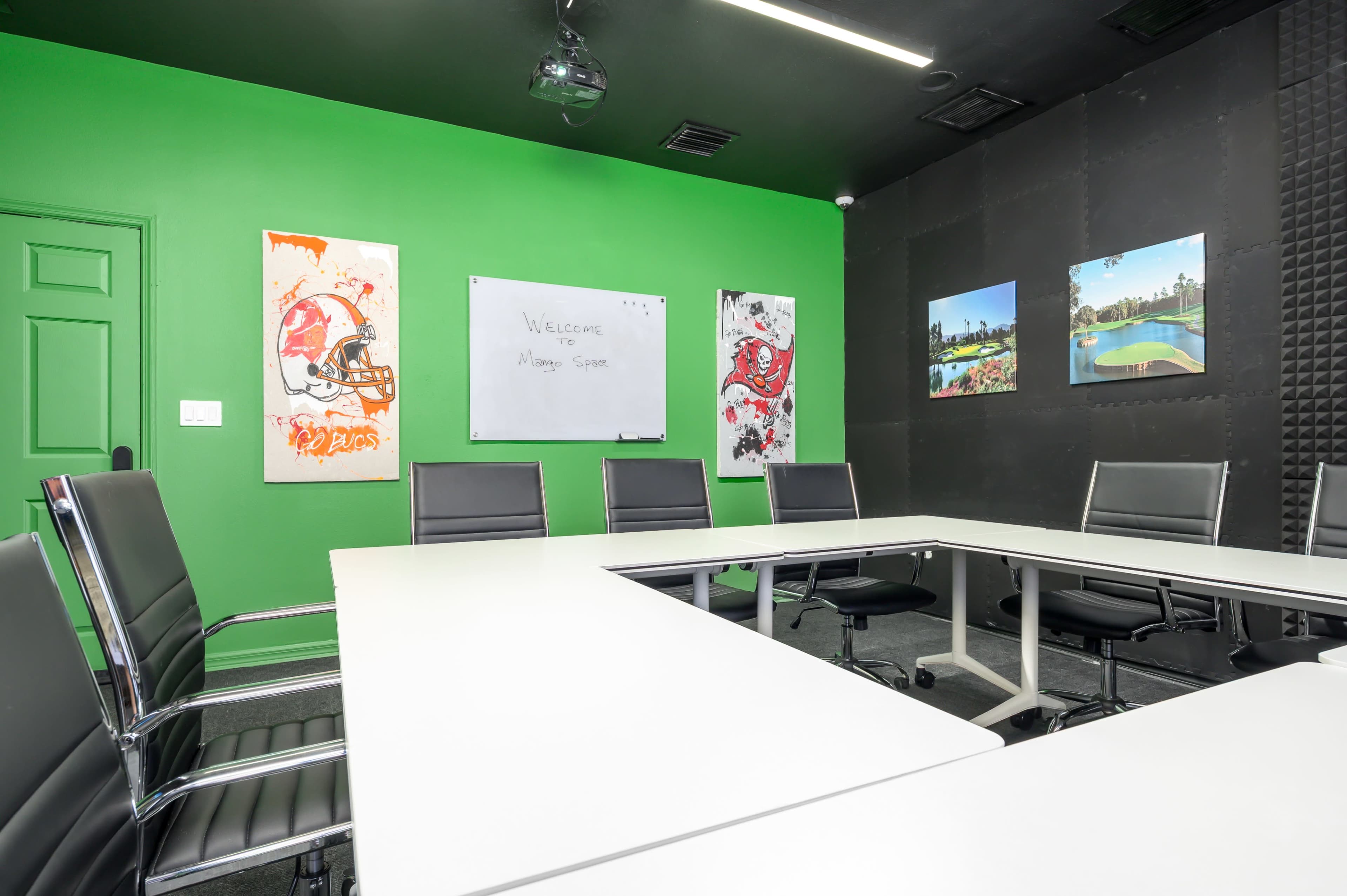 The image shows a modern conference room with a green accent wall, a whiteboard, and black and white artwork displayed on the walls.