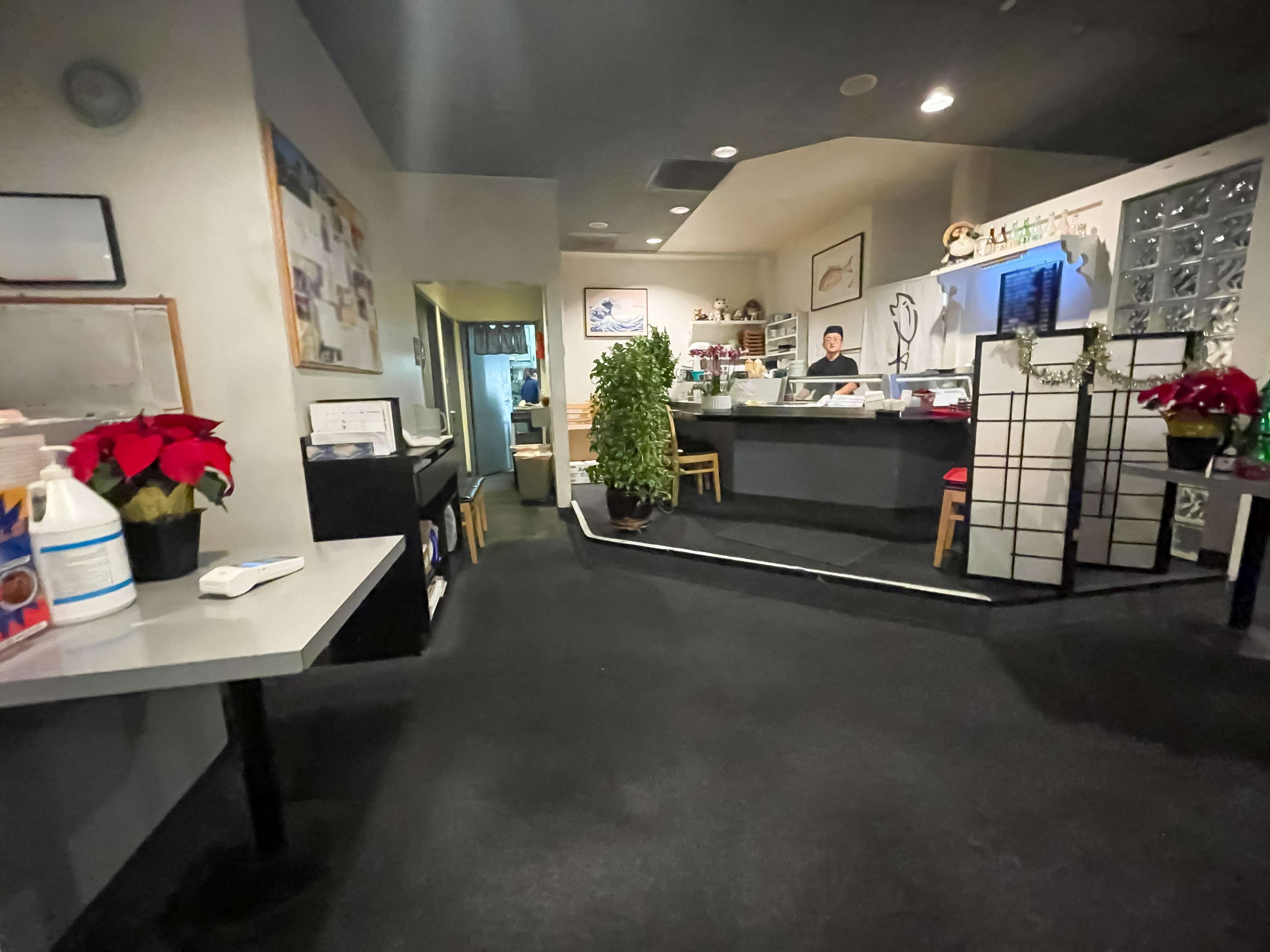 The image shows the interior of a restaurant with a reception desk, tables, and decorative plants.