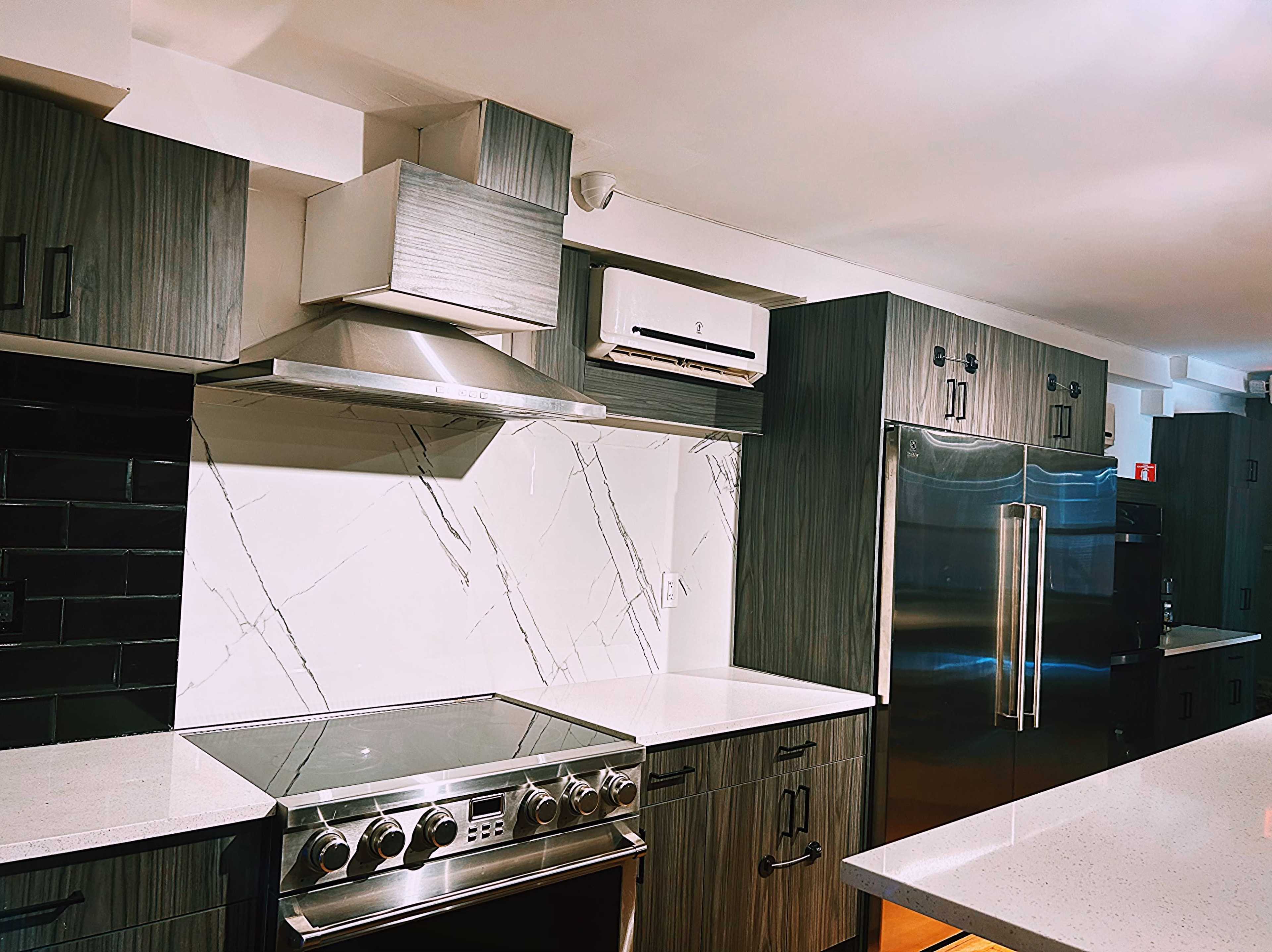 The image shows a modern kitchen featuring dark cabinetry, a stainless steel refrigerator, a built-in oven, and a marble-patterned backsplash.