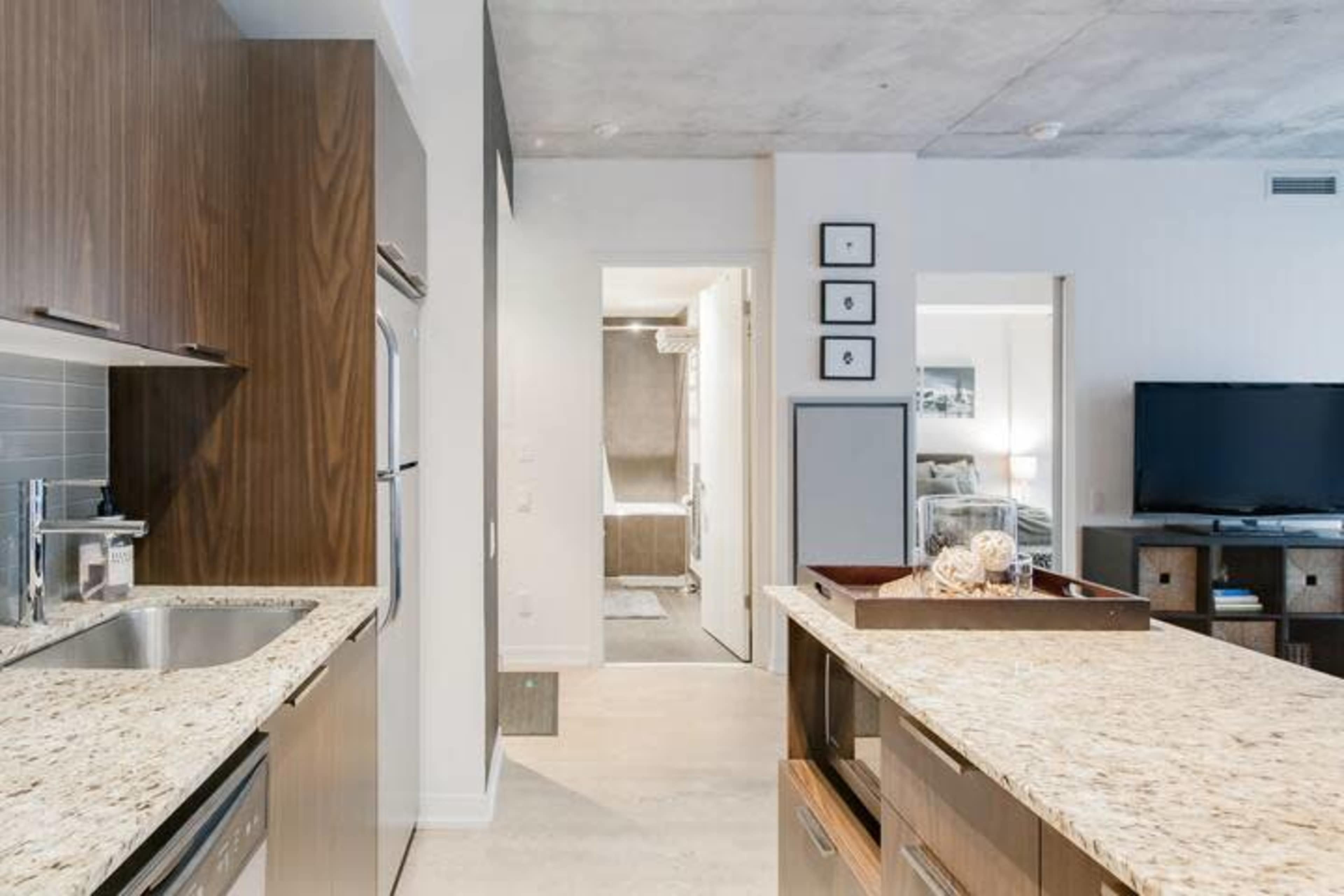 A modern kitchen with granite countertops leads to a hallway and a living area featuring a television and a bedroom door.