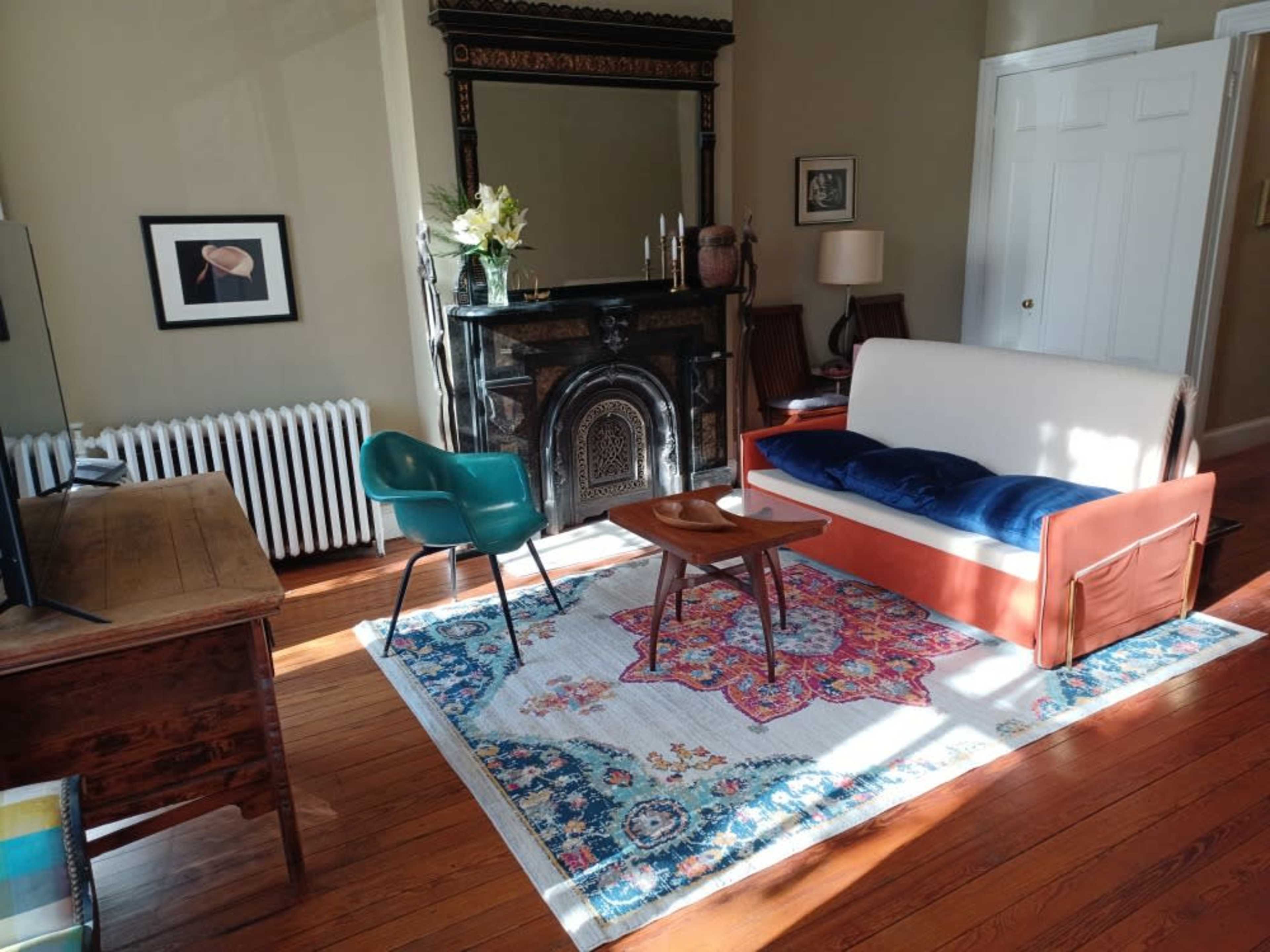 The image shows a cozy living room featuring a white and orange sofa, a green chair, a wooden table, and a decorative rug in front of a radiator and a black fireplace.