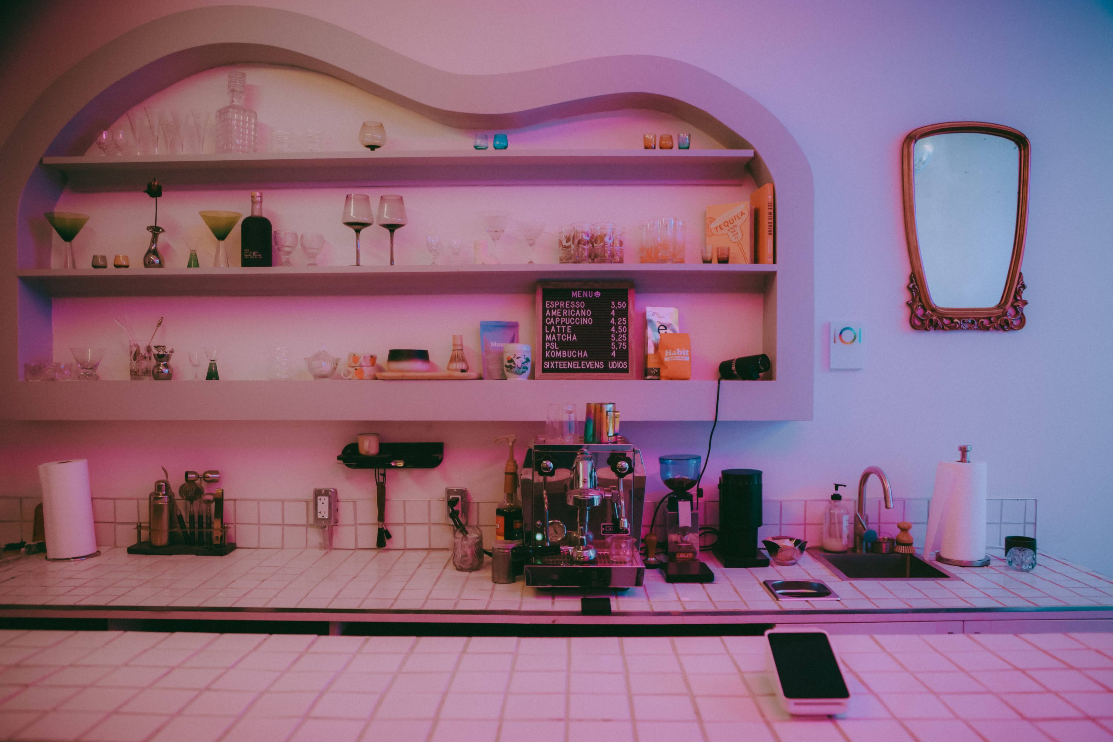 A modern kitchen bar features a variety of glassware on shelves above a coffee machine and countertop, with a mirror and a smartphone placed on the surface.