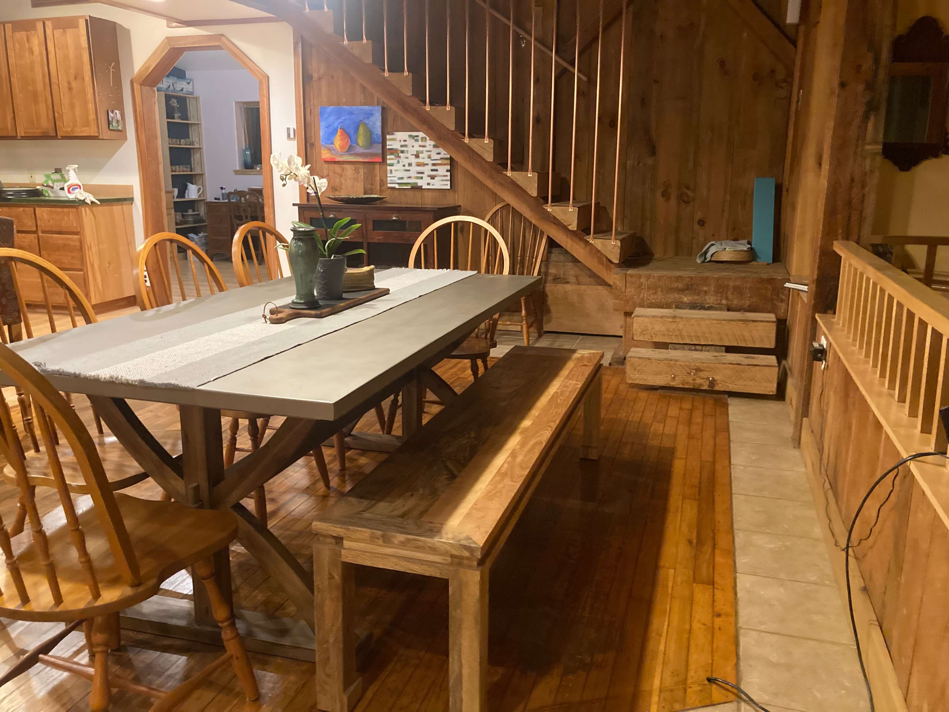 A wooden dining area with a large rectangular table and benches, situated next to a staircase and a wall featuring wooden paneling.