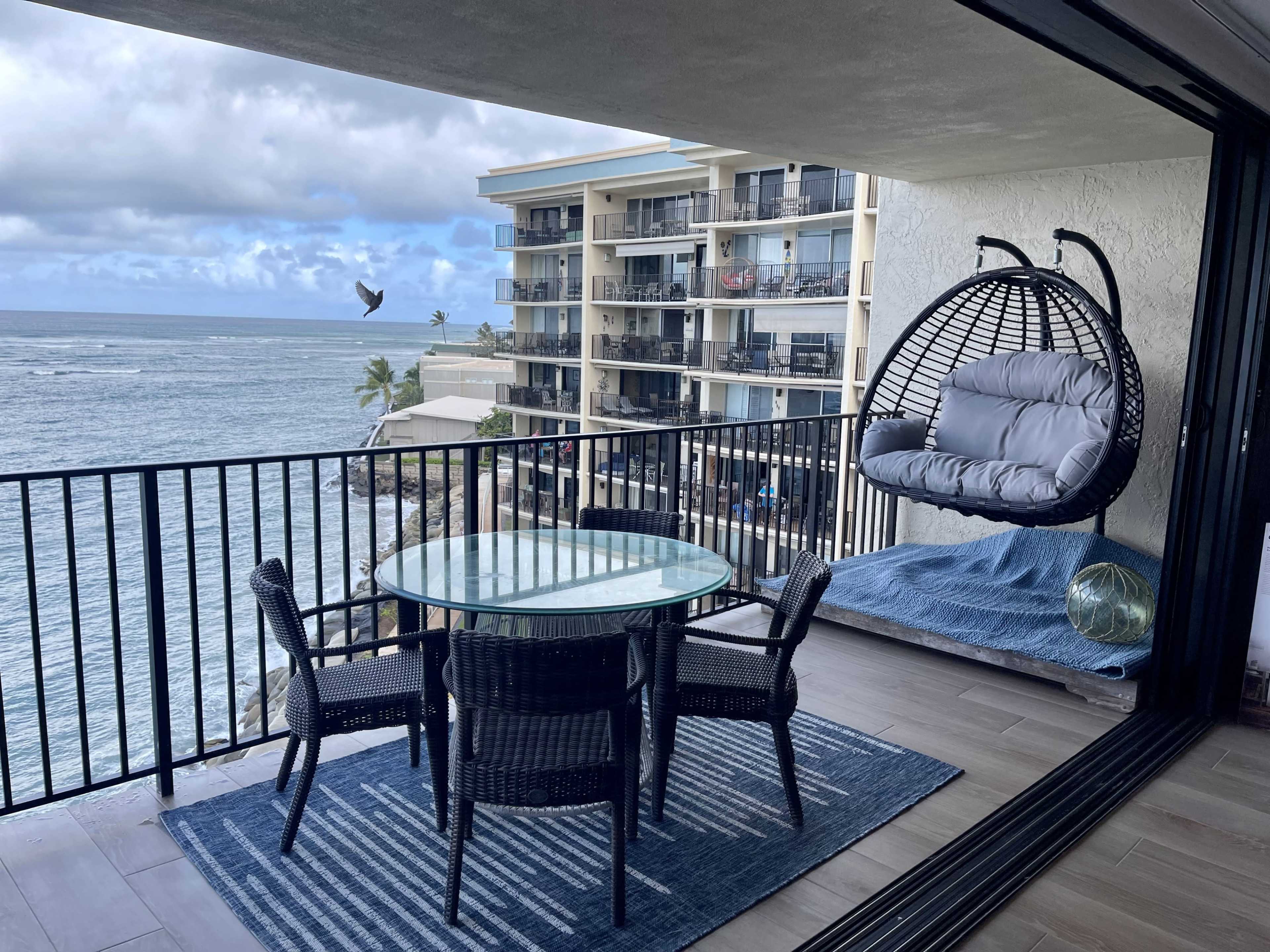 A balcony featuring a glass table with four wicker chairs, a hanging chair, and an adjacent view of the ocean and nearby buildings.