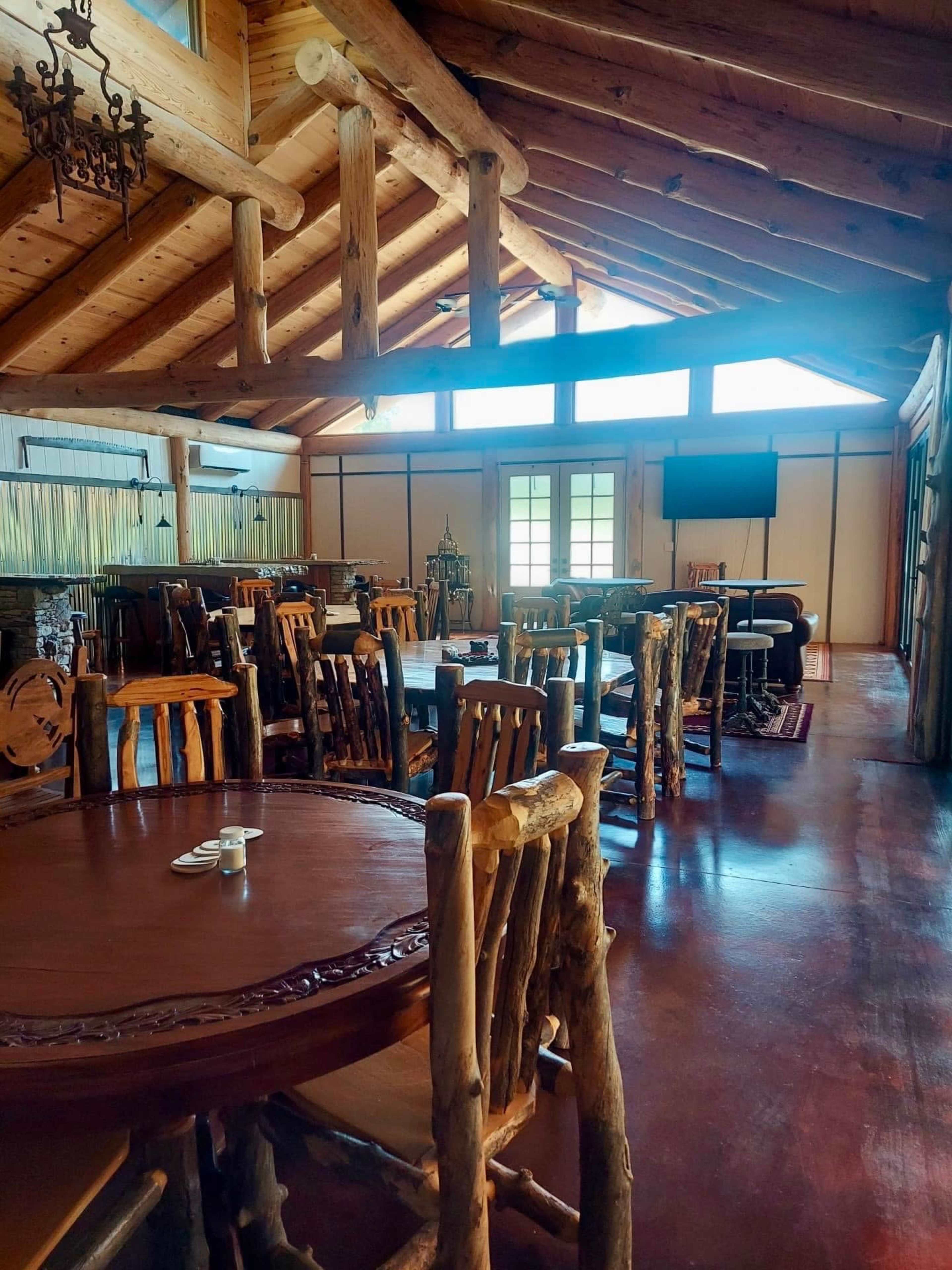 The image shows a rustic dining area with wooden tables and chairs, featuring high ceilings supported by exposed beams.