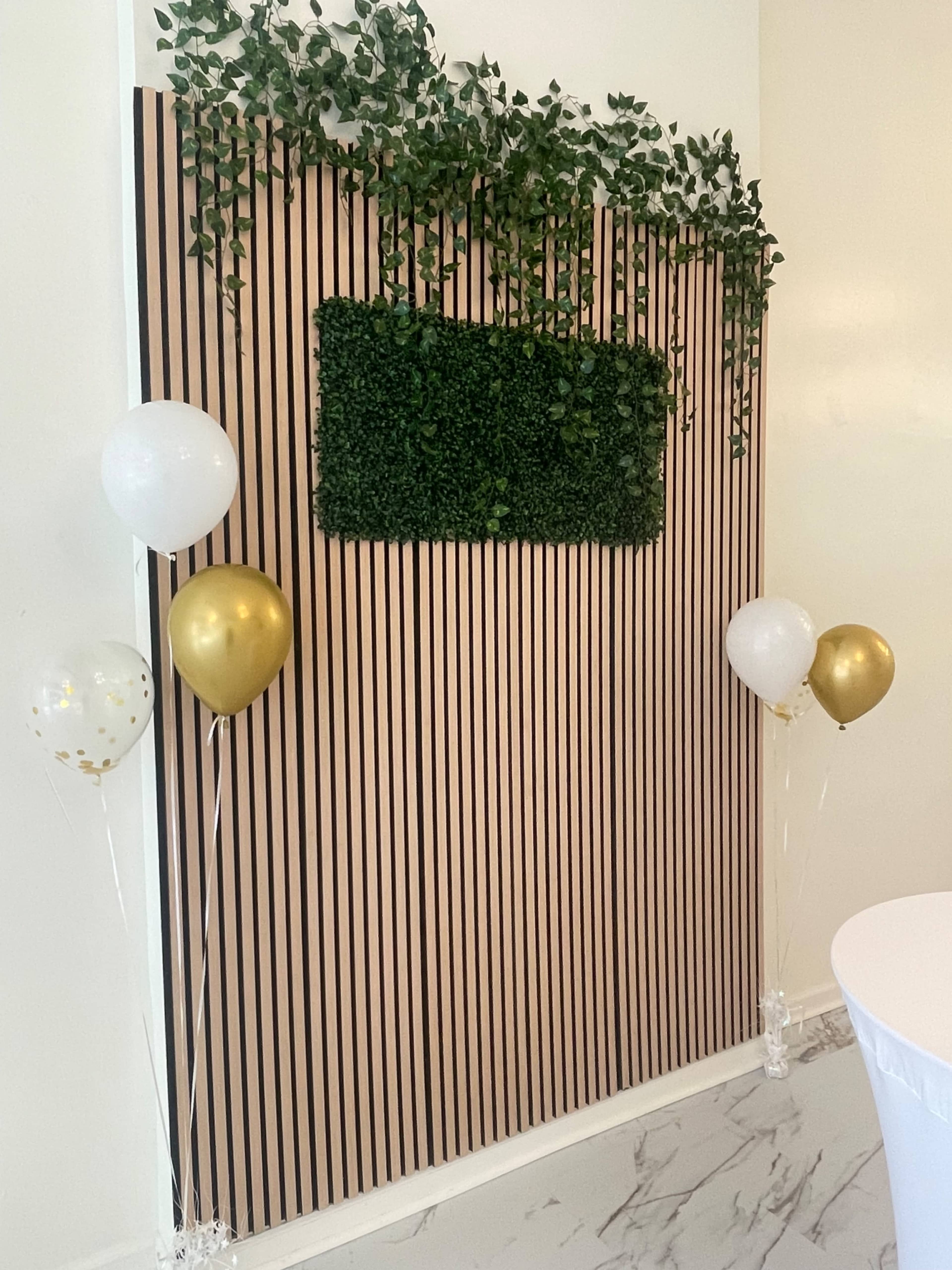 The image shows a vertical backdrop made of alternating striped panels with a section of artificial greenery at the center, accompanied by balloons in gold and white on either side.