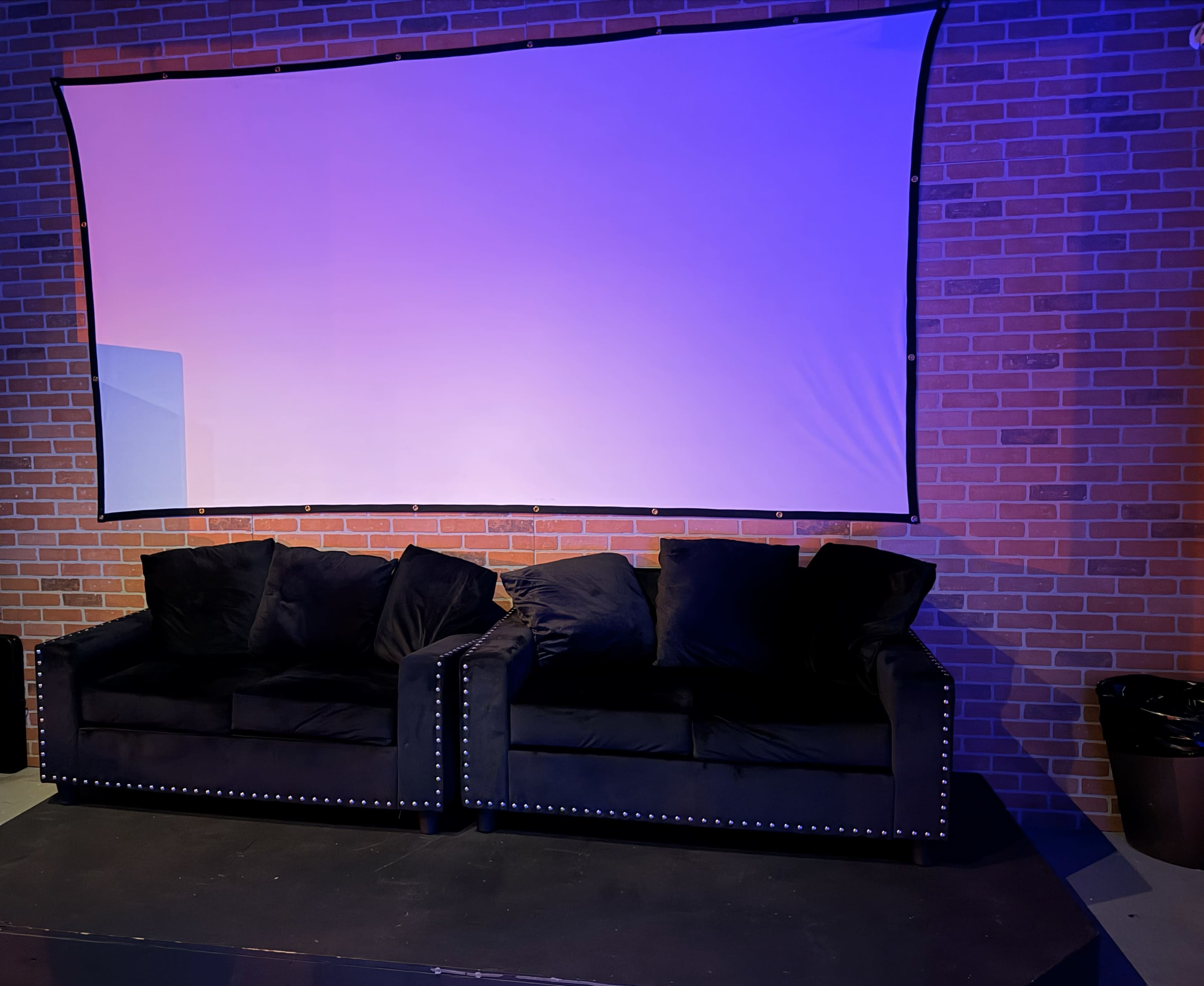 Two black couches with decorative studs are positioned in front of a blank movie screen against a brick wall.