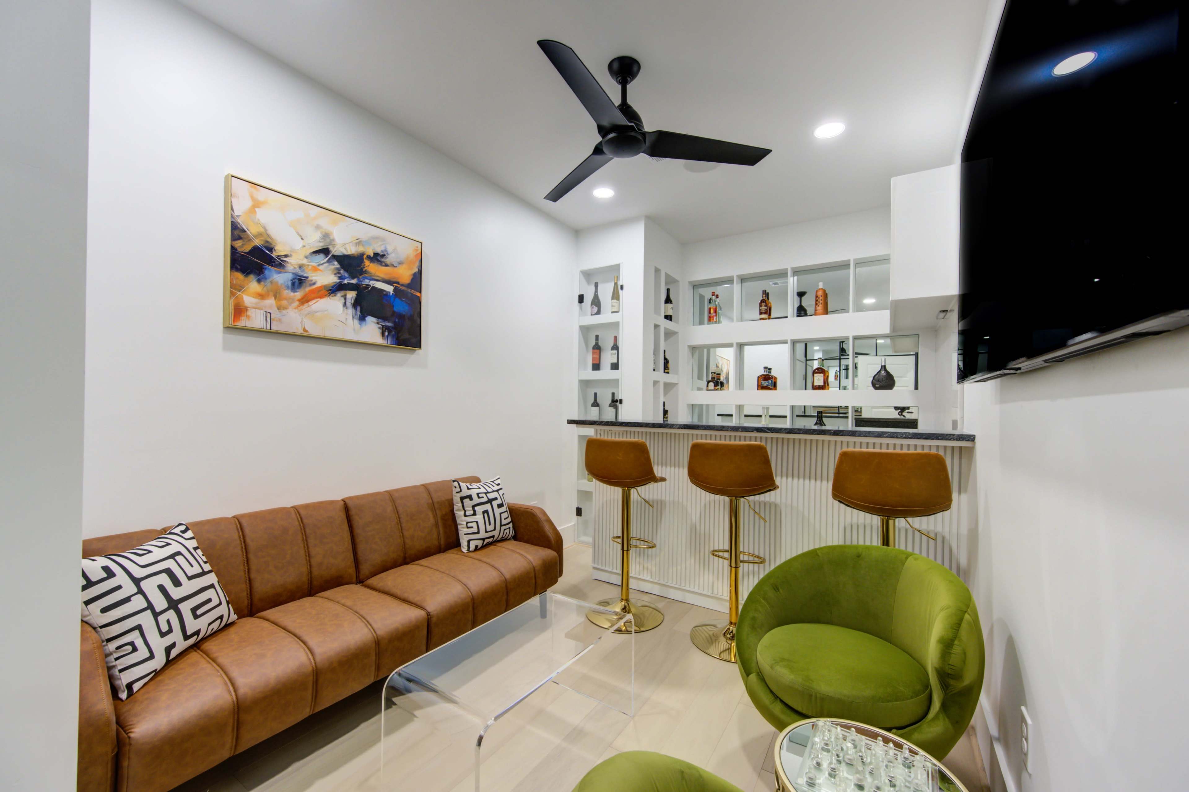 A modern lounge area with a brown sofa, two green chairs, and a well-stocked glass bar in the background.
