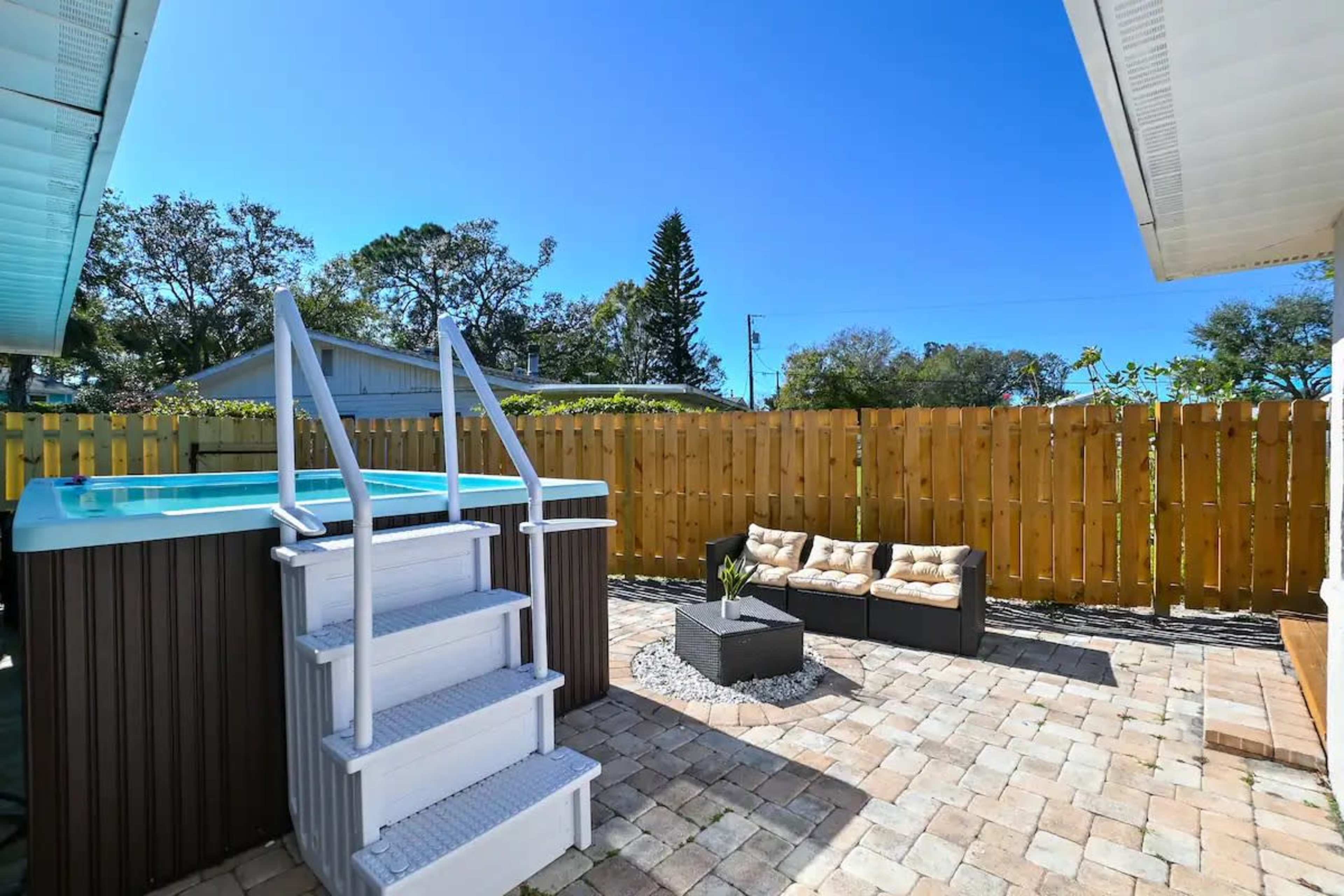 A small outdoor patio with a wooden fence, a raised pool, and a seating area made of black wicker furniture with cushions.