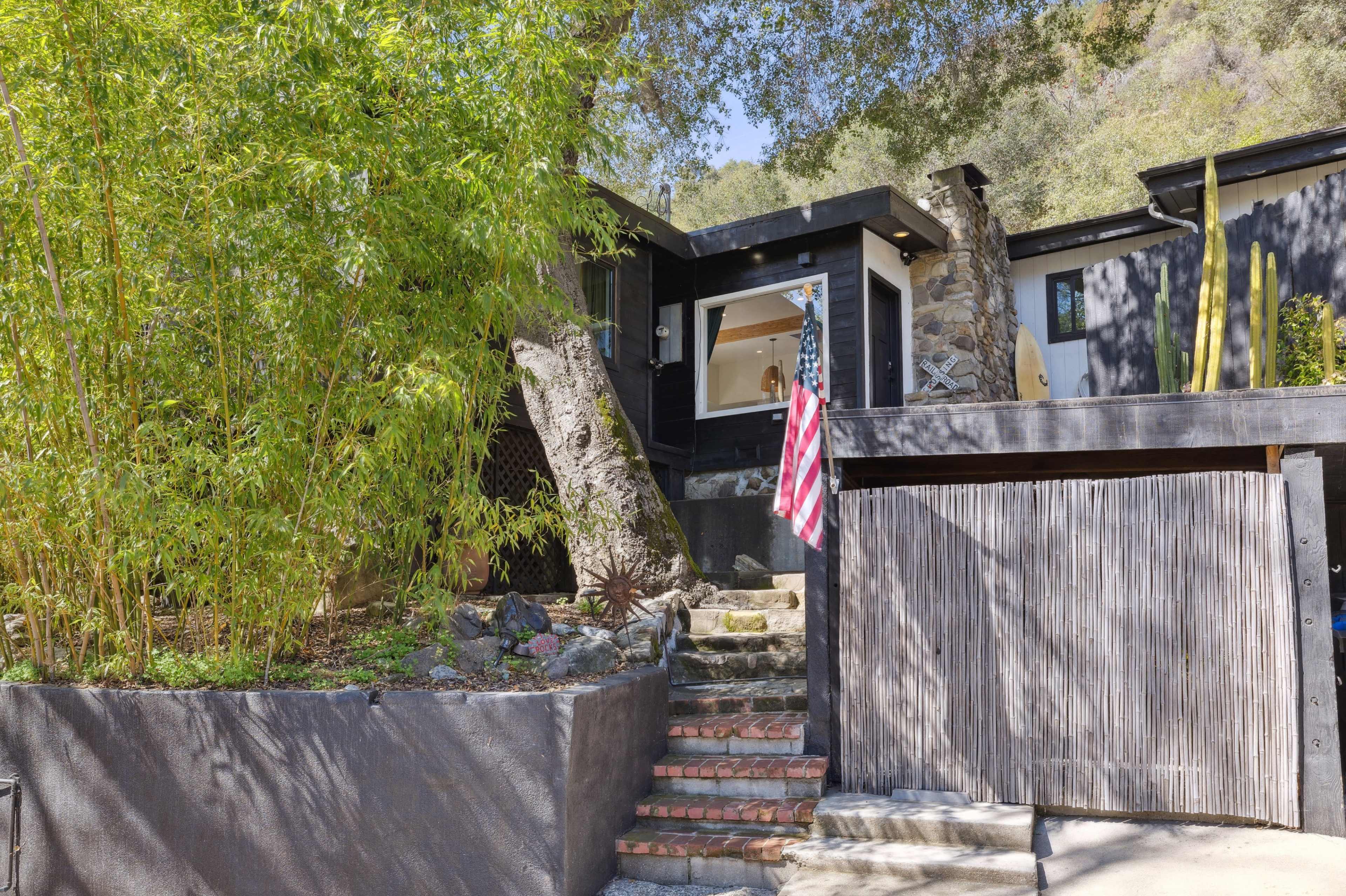The image shows a modern black house with stone accents, a wooden gate, and a set of stone steps leading up to the entrance, surrounded by greenery and a flag displayed on the side.