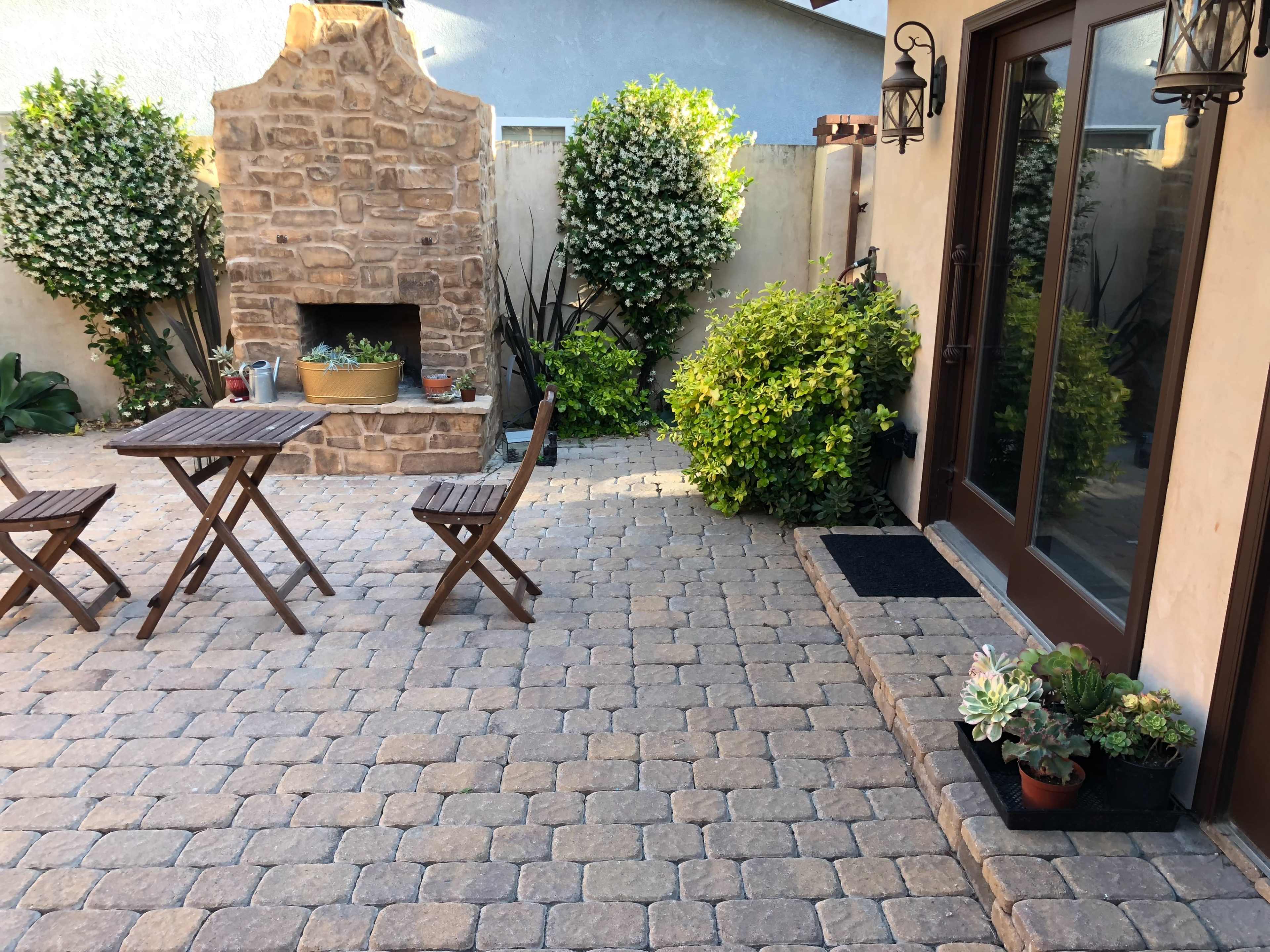 A stone patio features a small wooden table with two chairs, a decorative outdoor fireplace, and various plants along the walls.