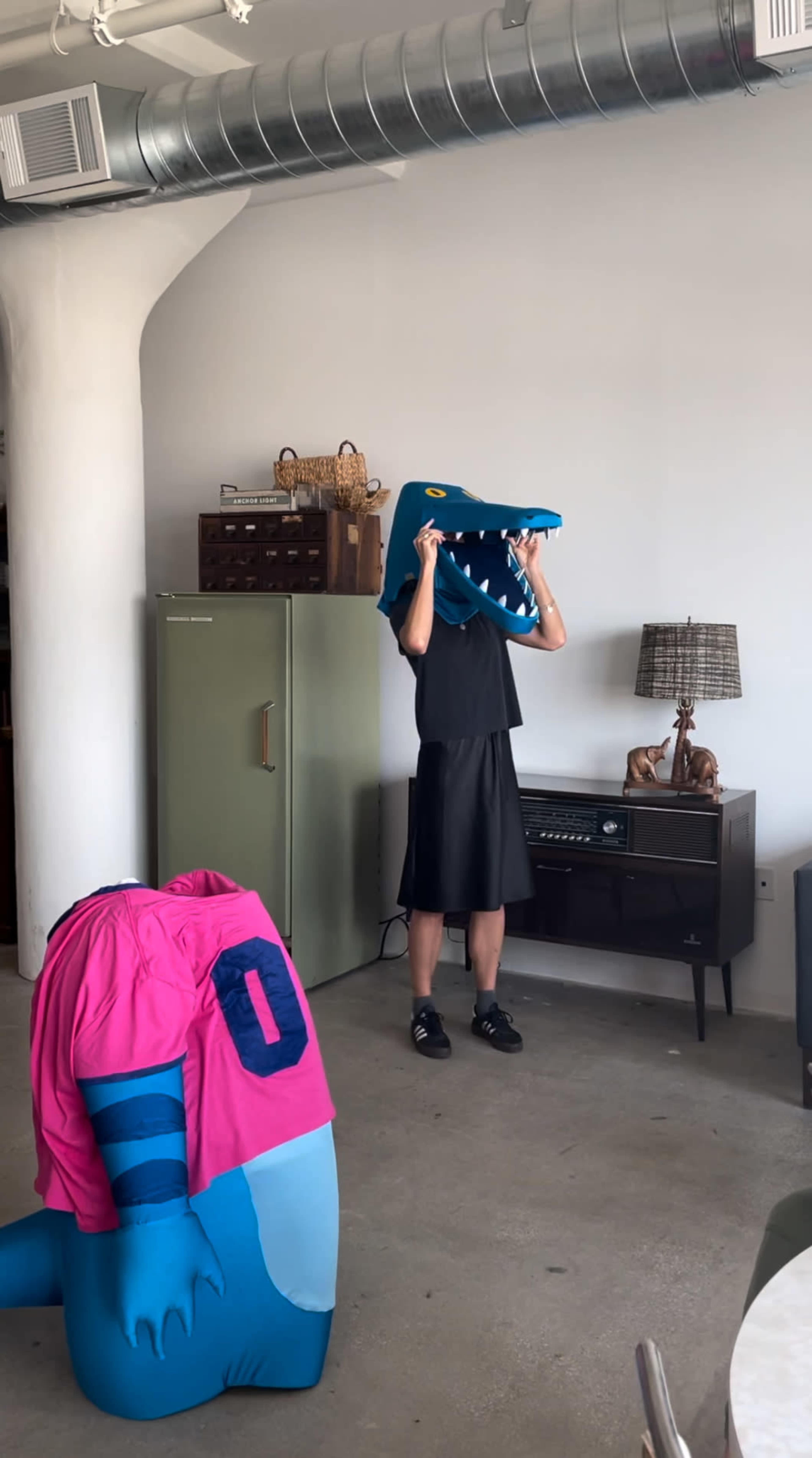 A person wearing a large dinosaur head costume stands in a minimalist room beside a pink dinosaur costume and a green refrigerator.