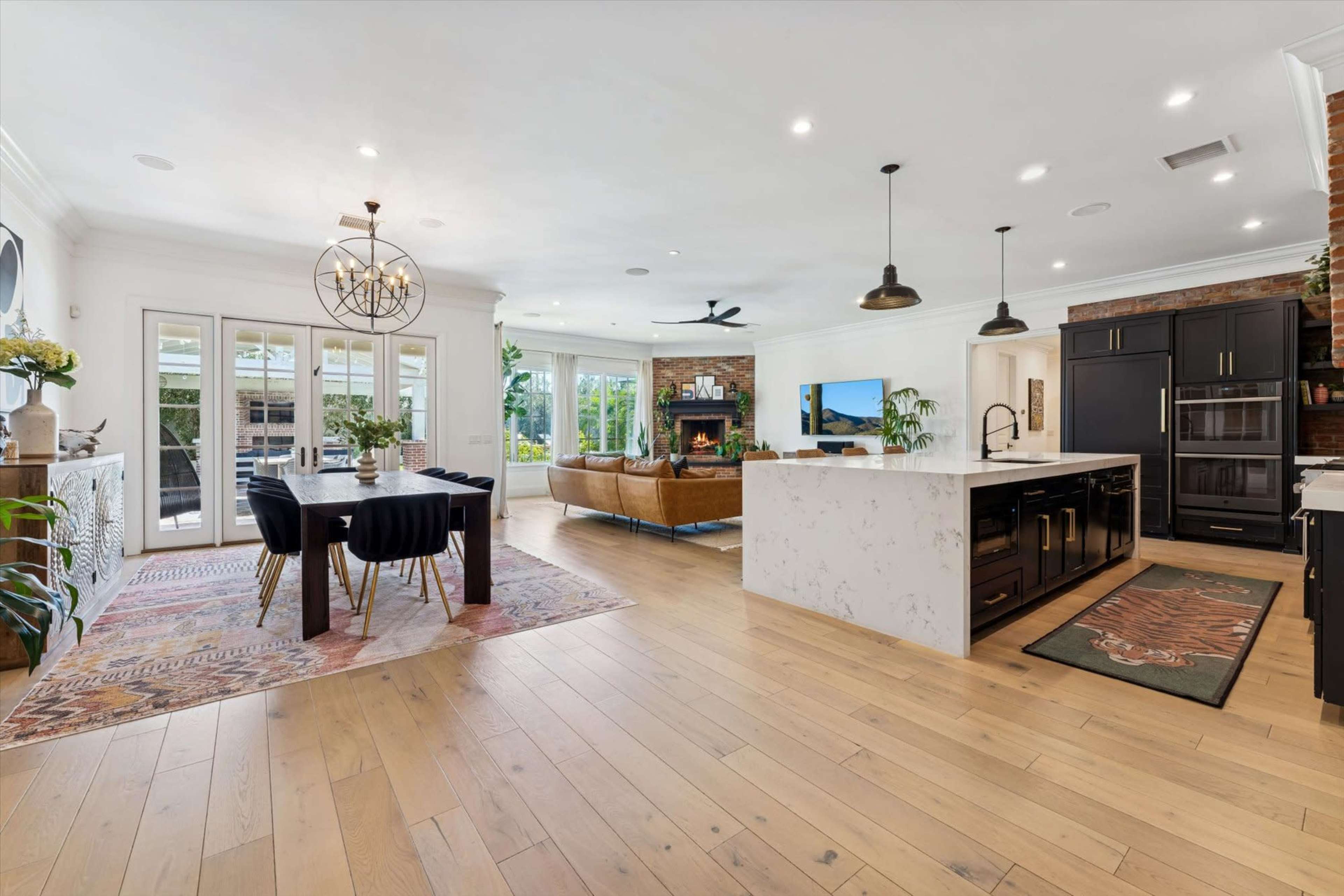 The image shows a modern kitchen and dining area featuring a large island, hardwood floors, a dining table, and a cozy seating area with a fireplace.