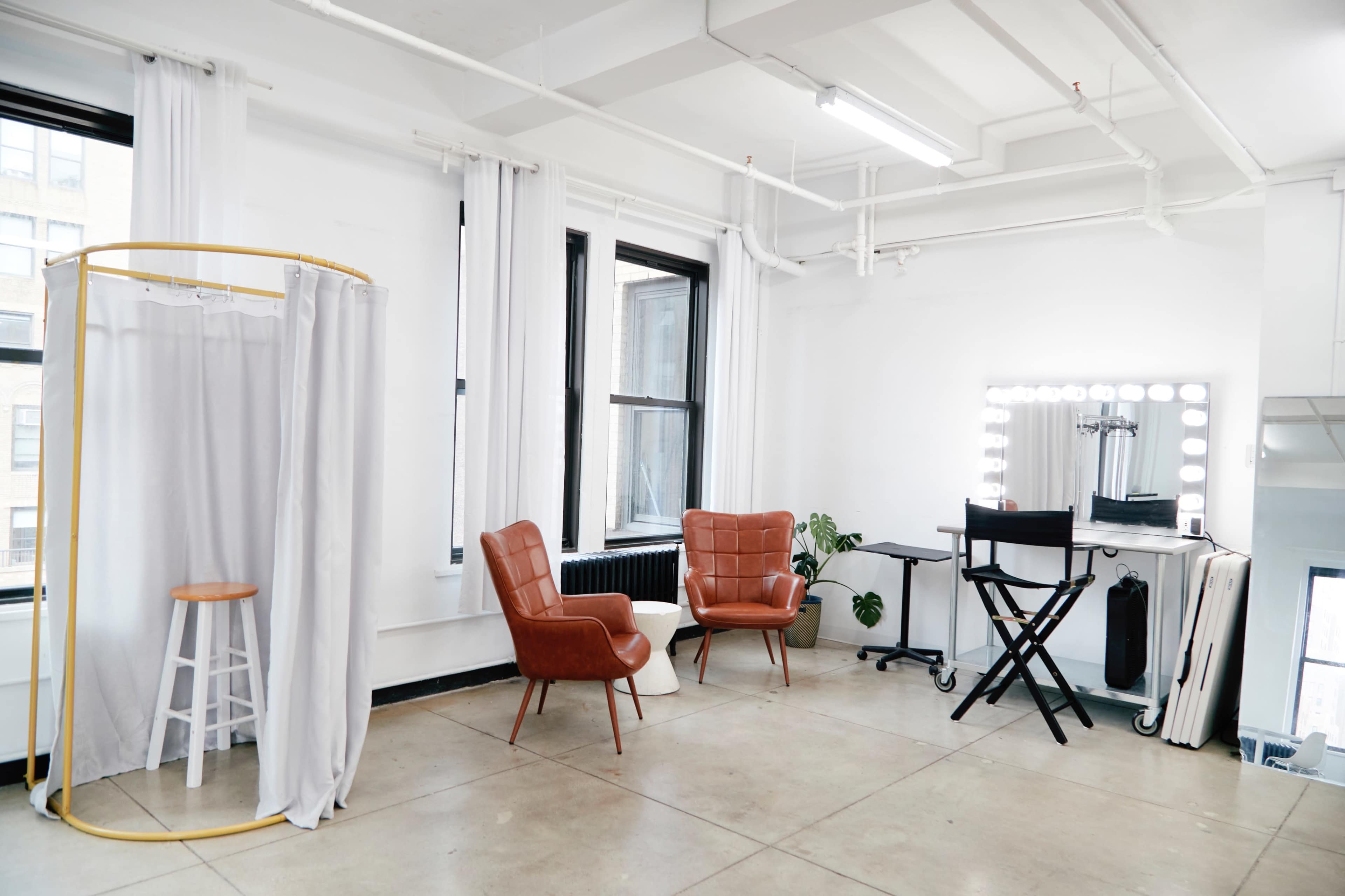 The image shows a bright, spacious room with two leather chairs, a small wooden stool, a portable dressing area with a curtain, a makeup station with lights, and large windows.