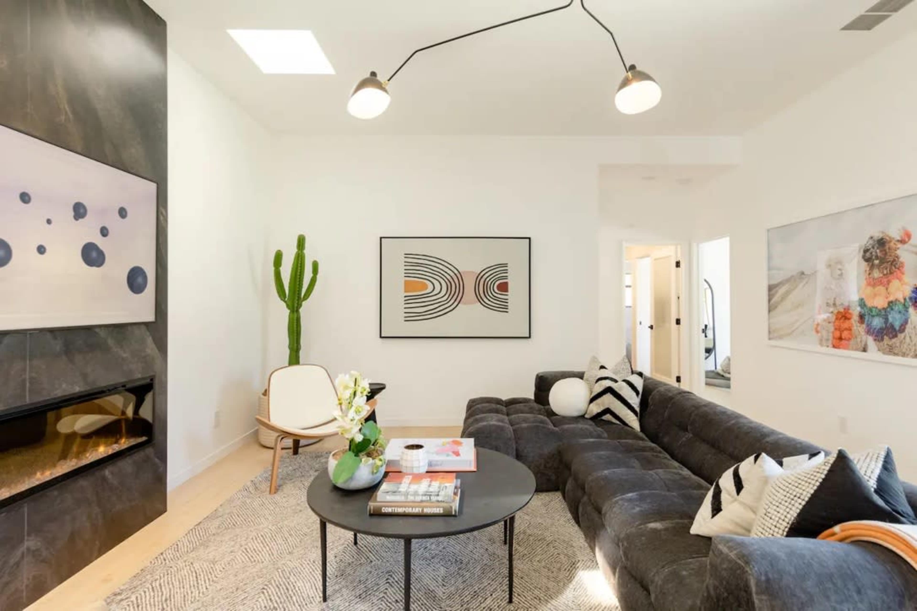 The image shows a modern living room featuring a dark sectional sofa, a round coffee table, a large wall-mounted television, and decorative art pieces, with a tall cactus in one corner.