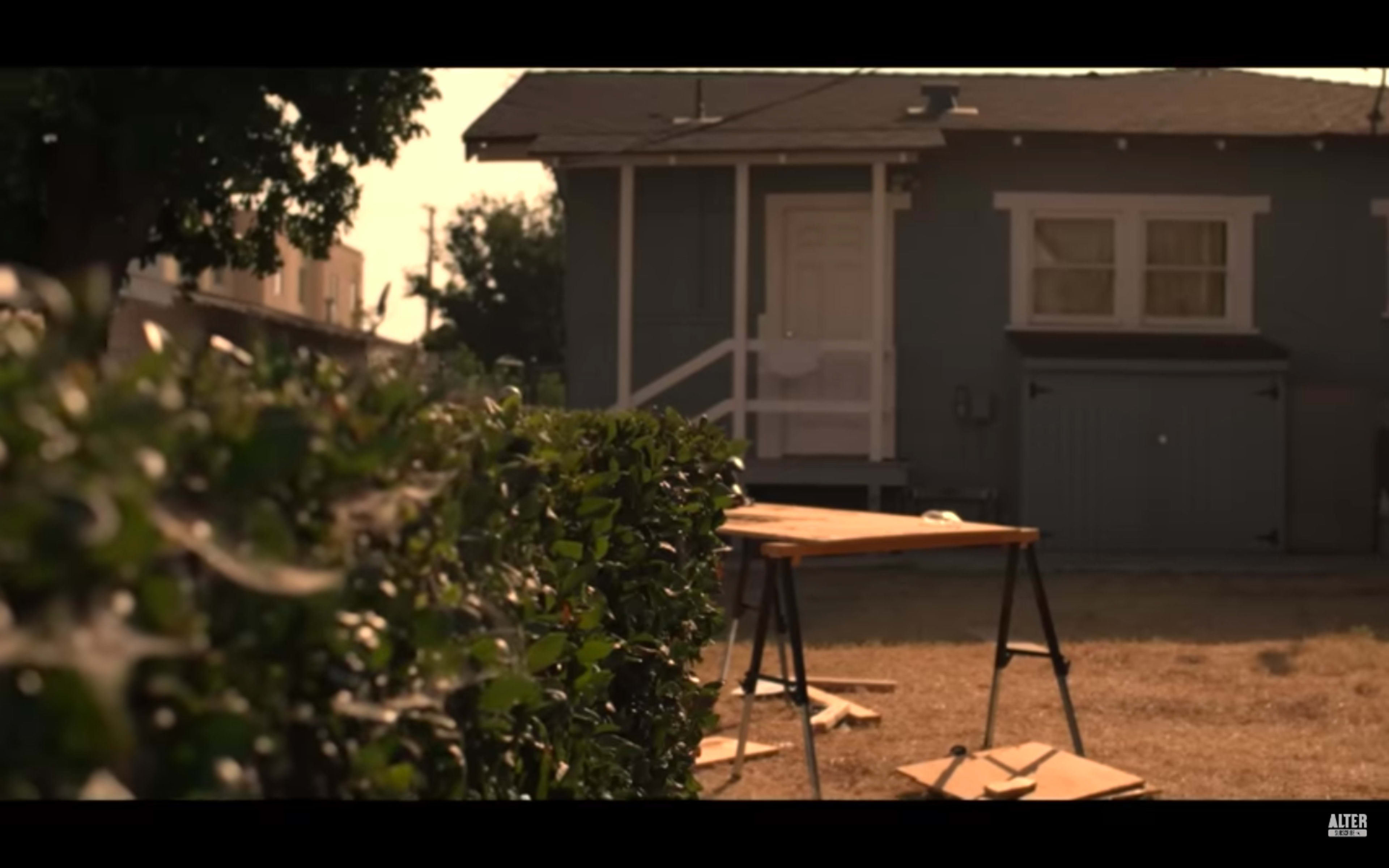 A wooden table stands on a patch of grass near a neatly trimmed hedge, with a light blue house and garage visible in the background.