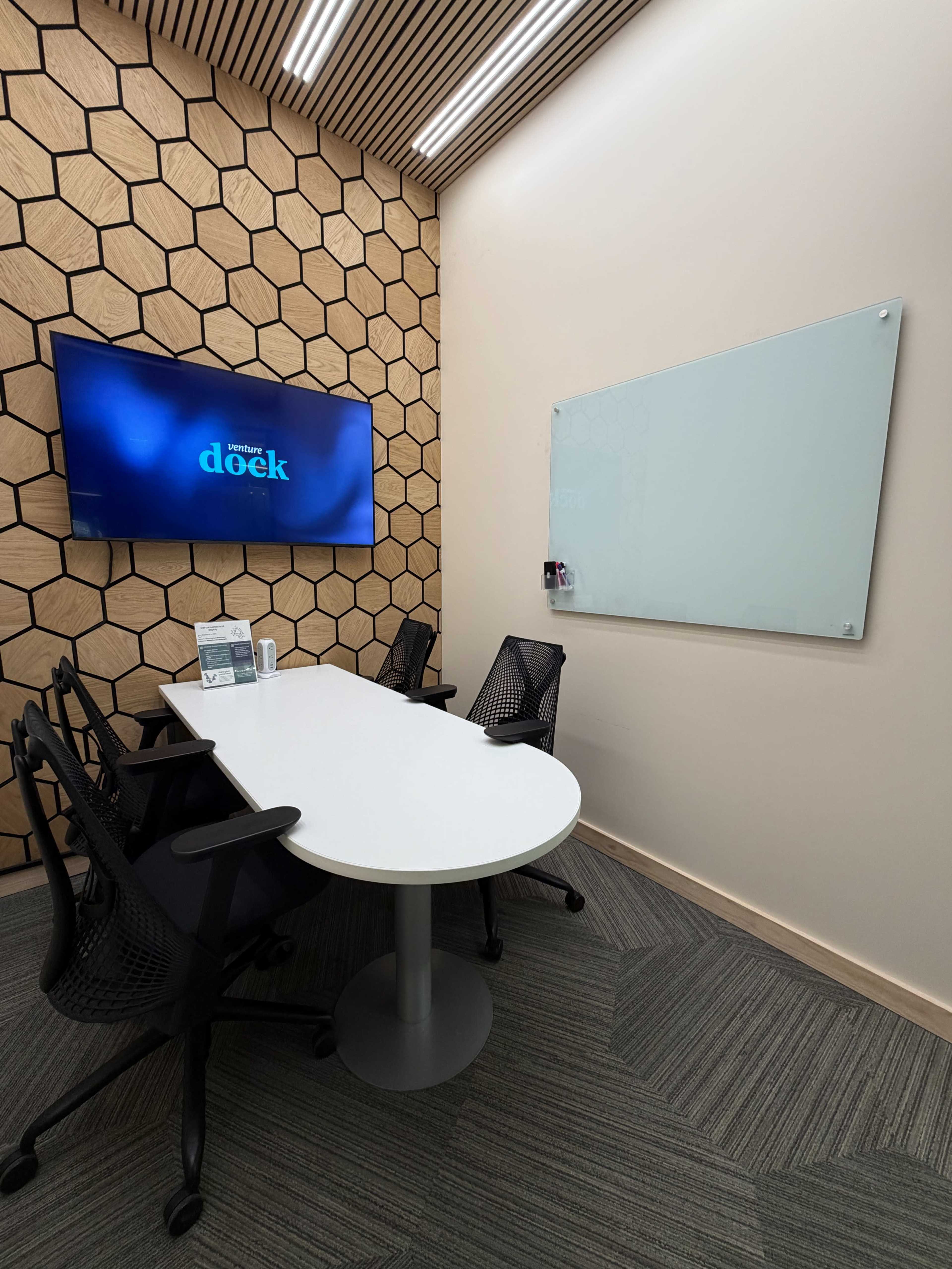 A modern conference room features a hexagon-patterned wall, a large screen displaying the word "doek," a whiteboard, and four black ergonomic chairs around a white table.