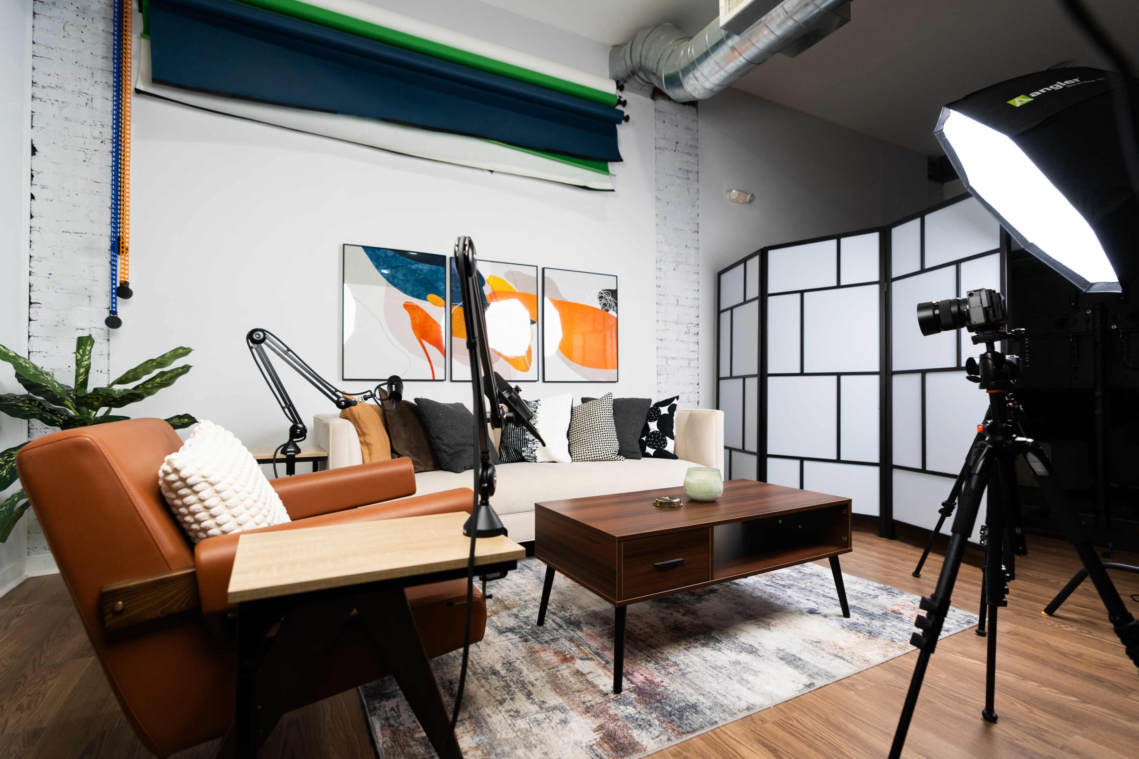 A modern living room setup with a sofa, various decorative cushions, a coffee table, a plant, and a camera on a tripod next to a softbox light.