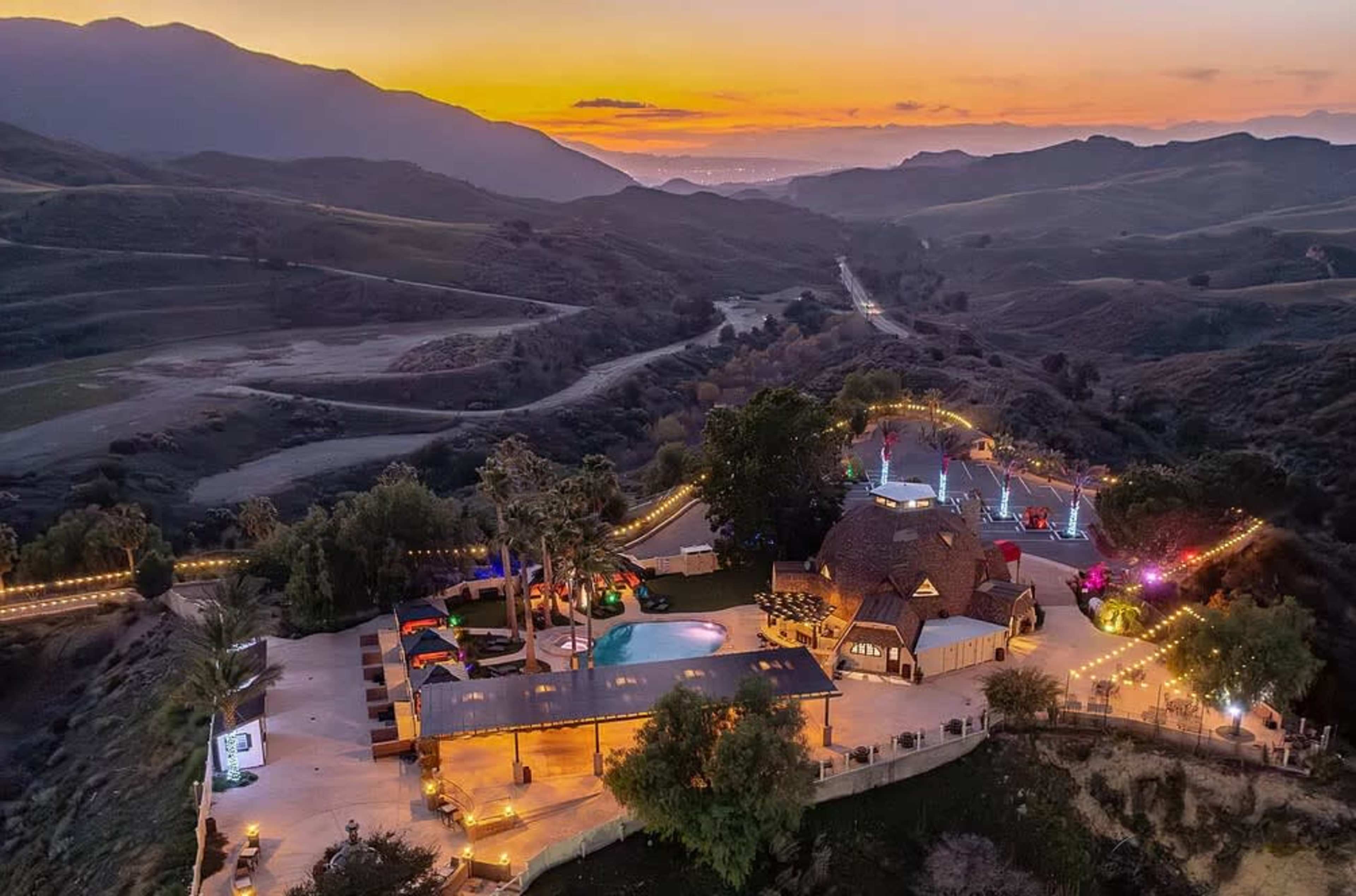 The image shows a scenic view of a large outdoor space with a pool and a house, set against a backdrop of rolling hills at sunset.
