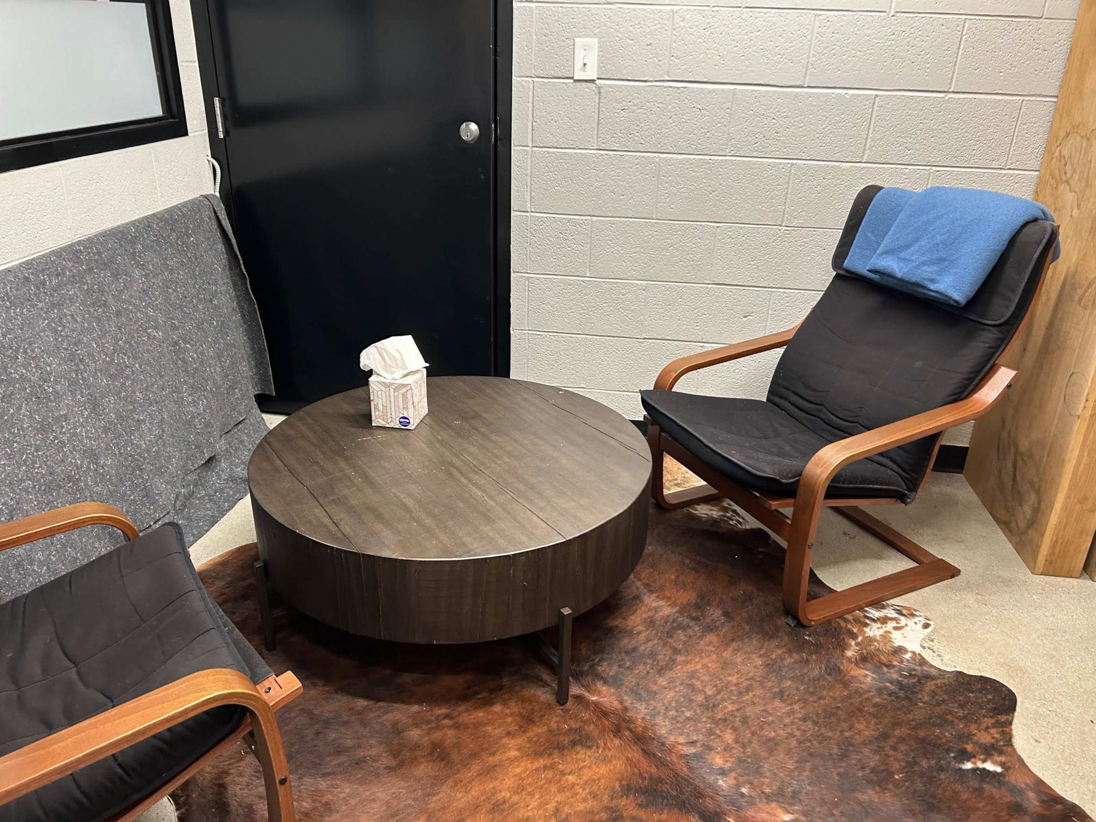 A small seating area with two black chairs, a round wooden coffee table, and a tissue box on top, set against a concrete wall.