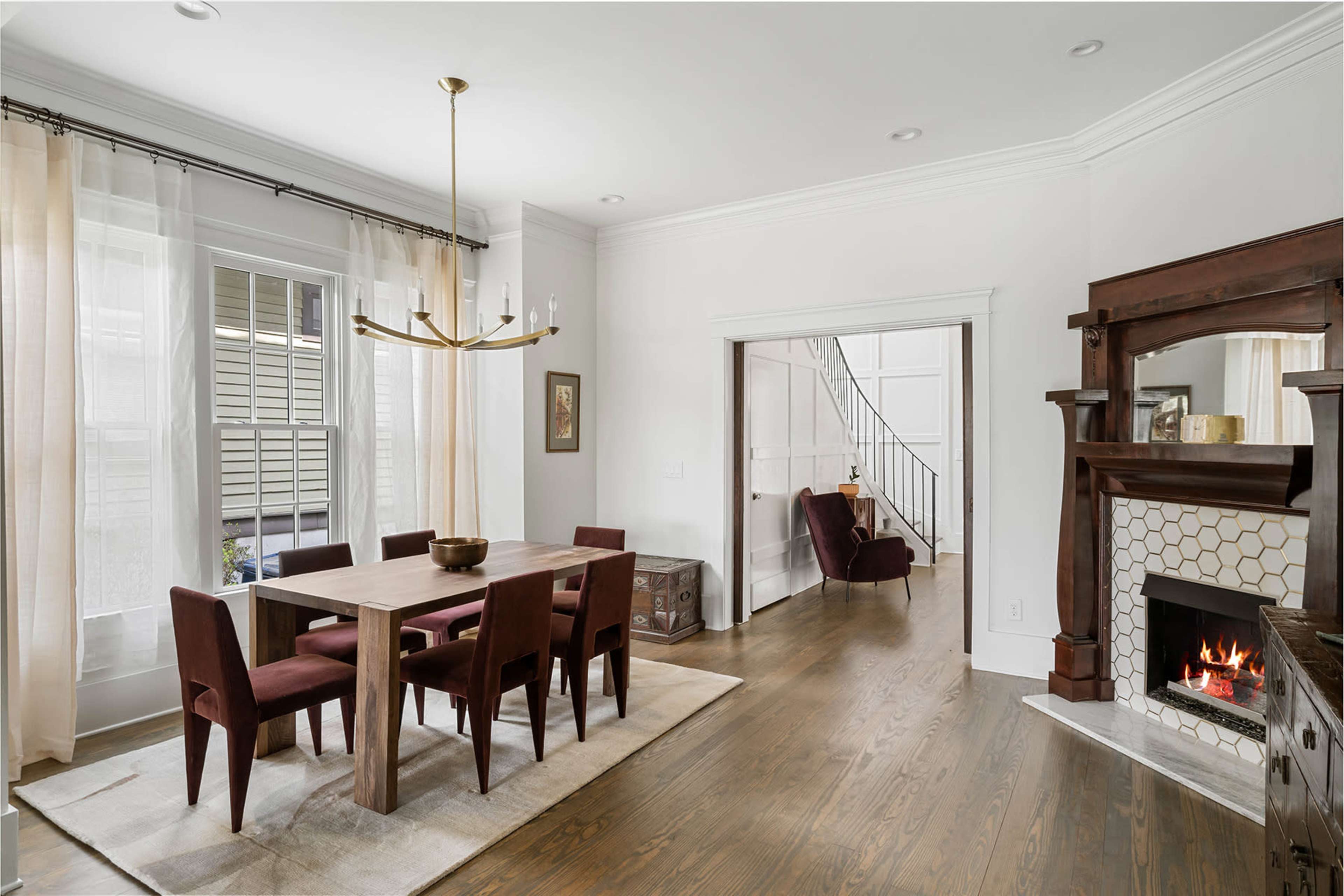 A dining area features a wooden table surrounded by burgundy chairs, with a fireplace and a door leading to a staircase in the background.