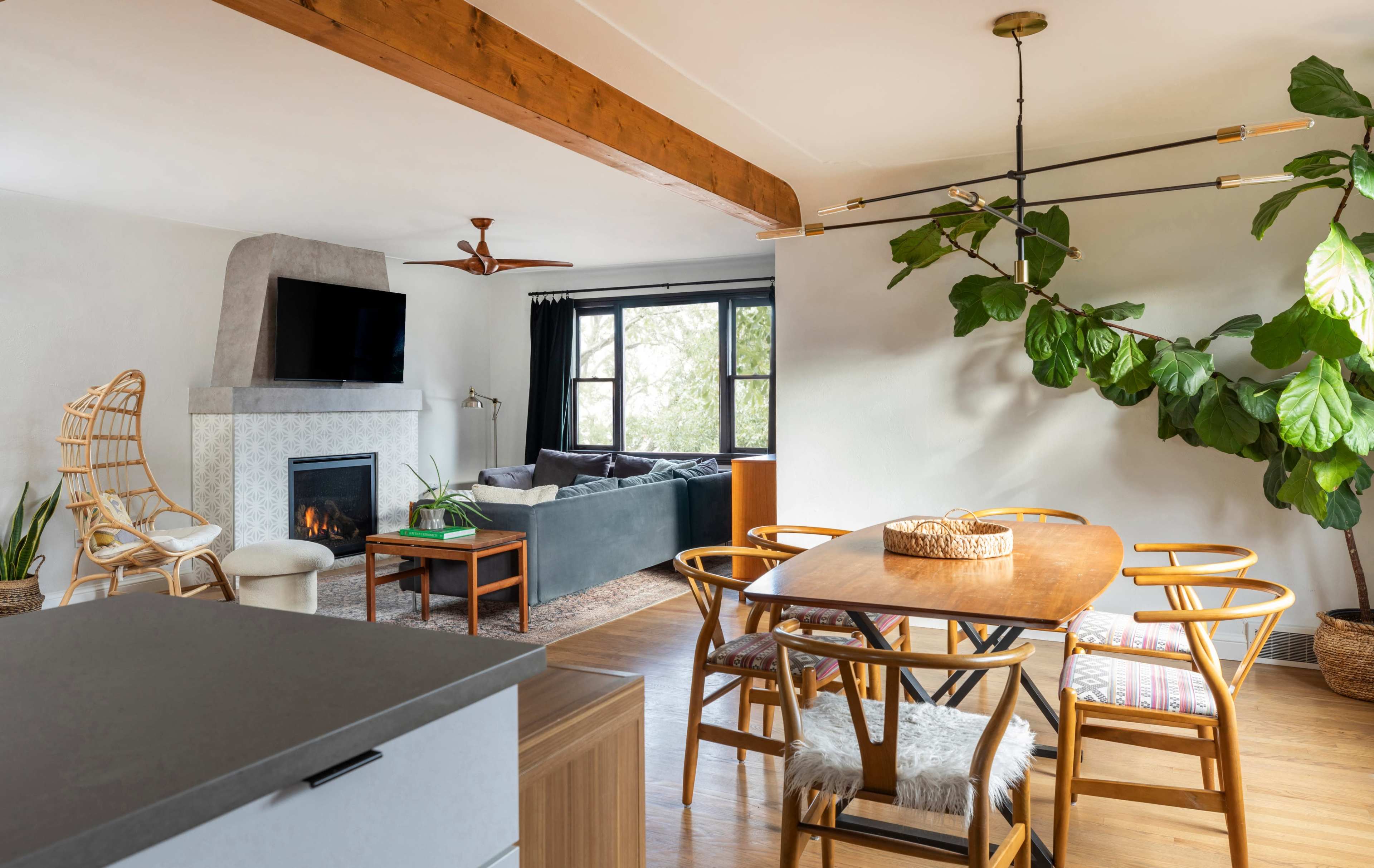 The image shows a modern living space featuring a dining area with a wooden table and chairs, a cozy seating area with a sofa, a fireplace, and a large window letting in natural light.