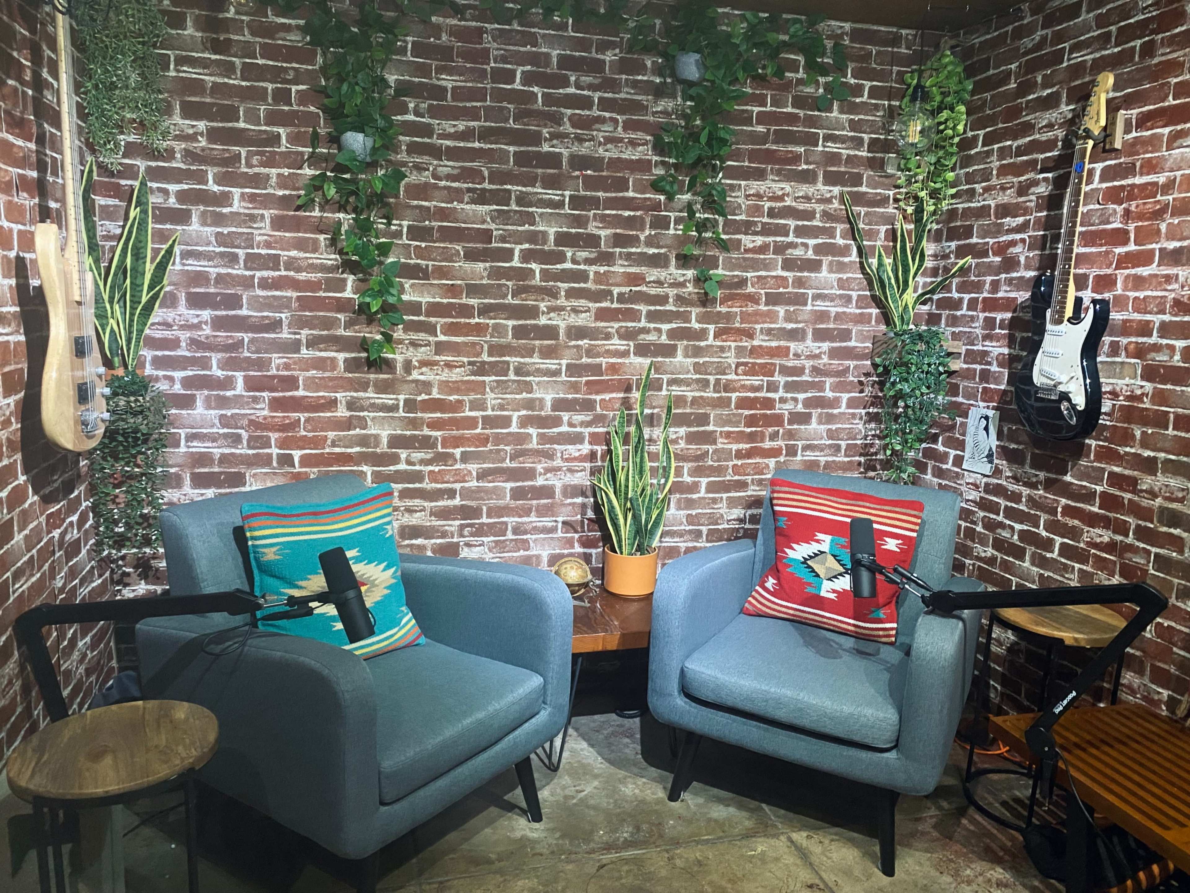 Two blue armchairs with colorful pillows positioned in front of a brick wall, accompanied by plants and a guitar on display.