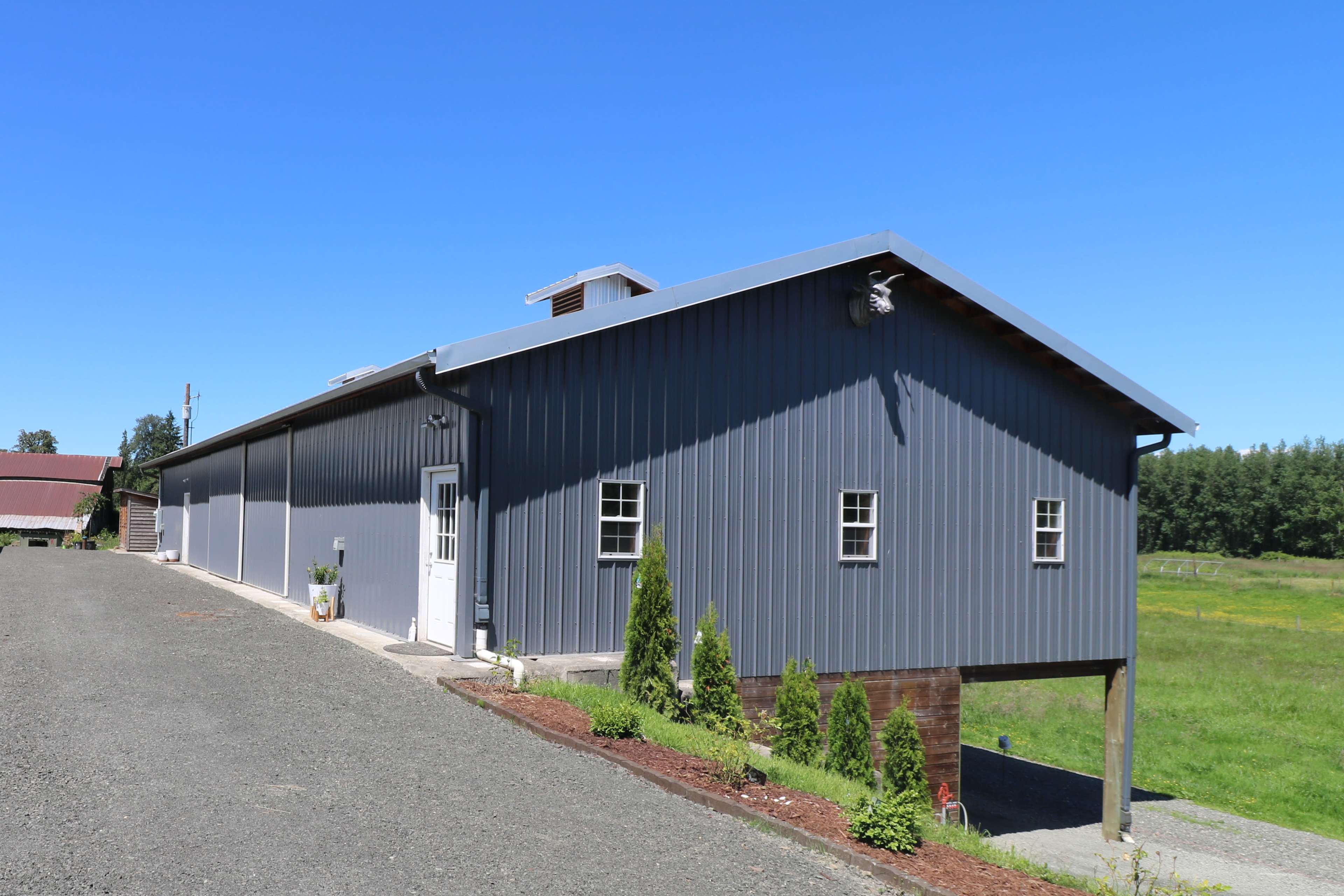 A metal-clad building with a gable roof is situated beside a gravel driveway, surrounded by grassy fields.