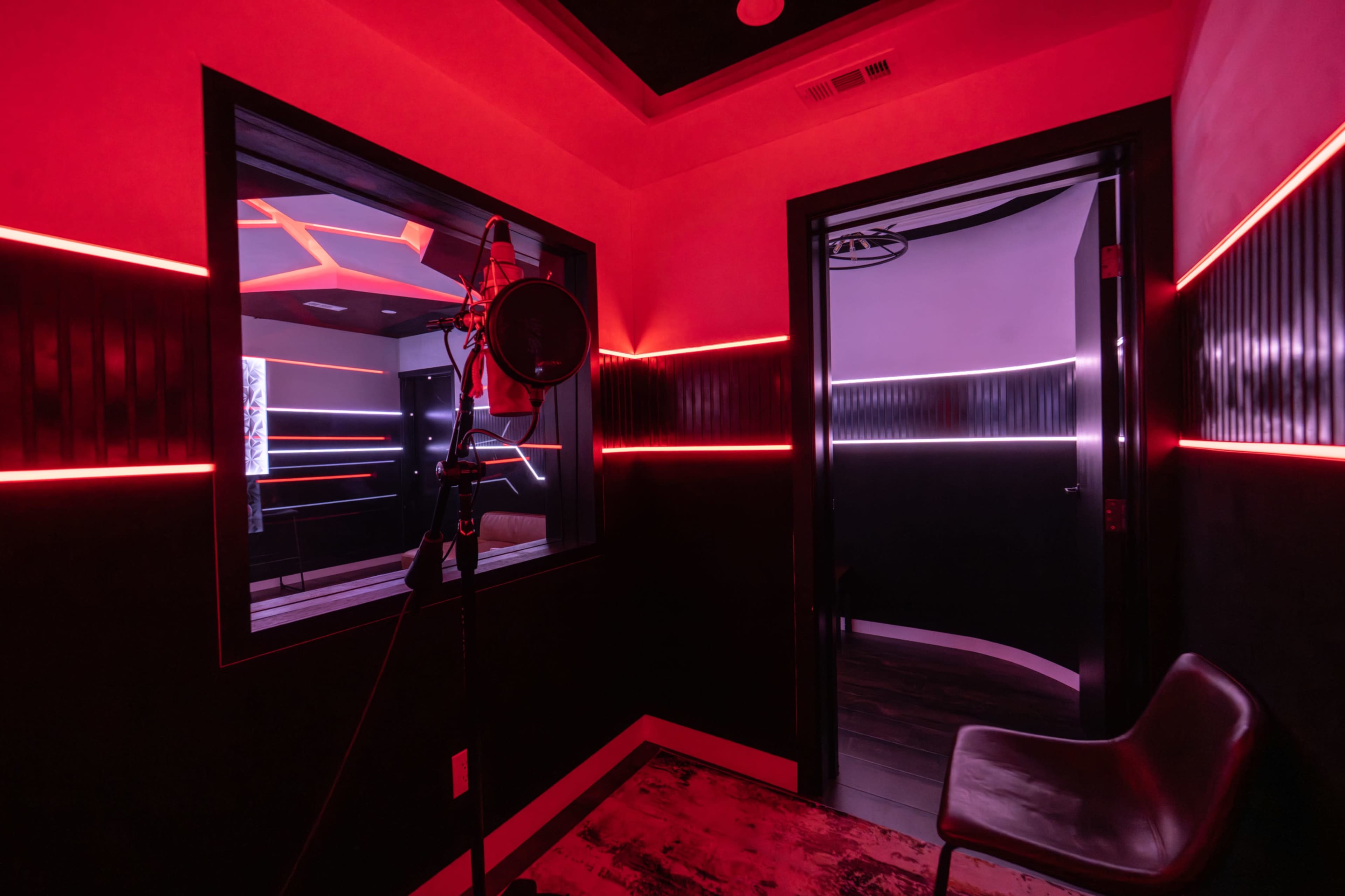 The image shows a modern recording studio with red ambient lighting, featuring a microphone on a stand and a glass window overlooking another room.