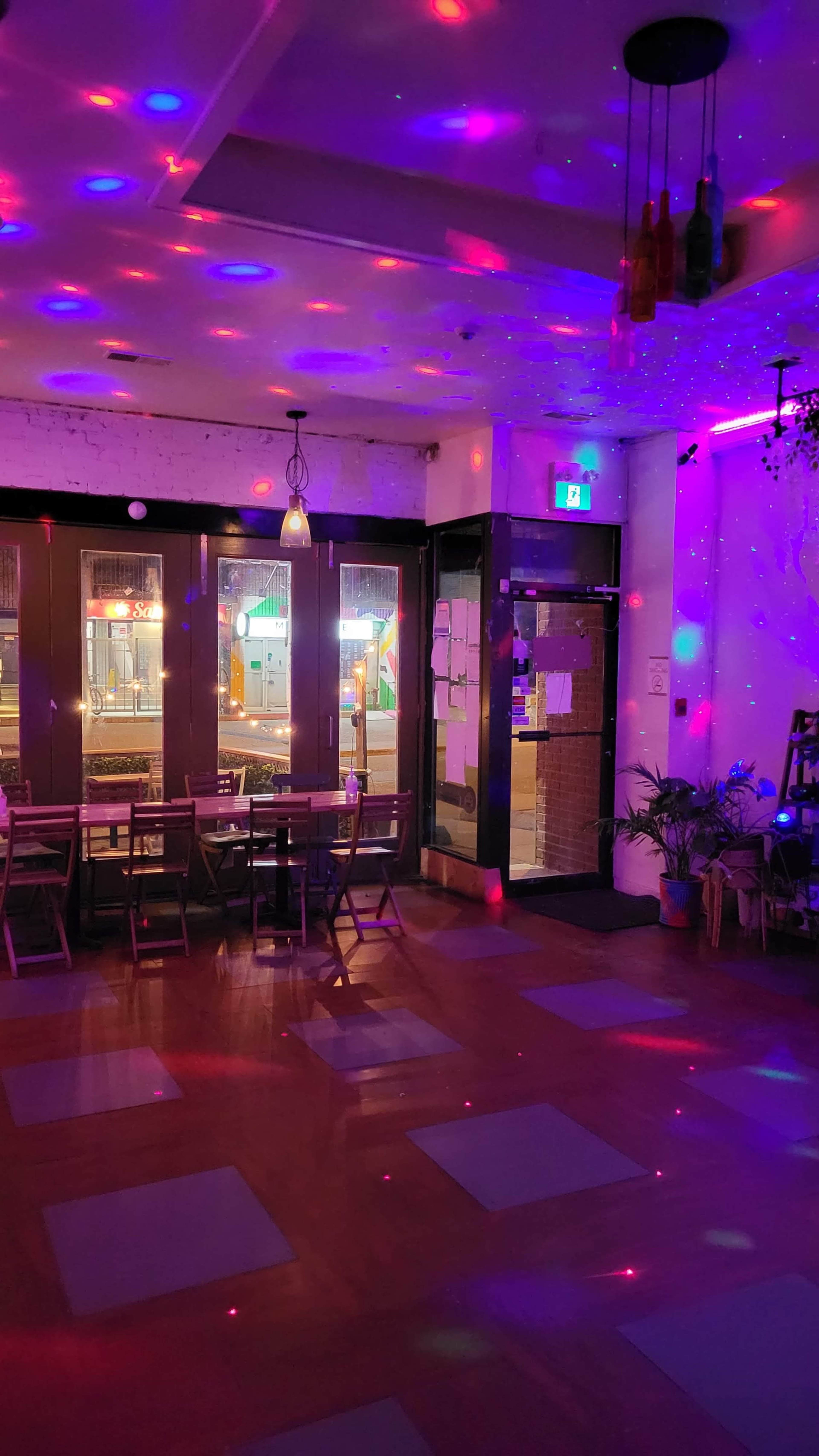 The interior of a dimly lit venue features tables and chairs with colorful lights casting patterns on the floor.
