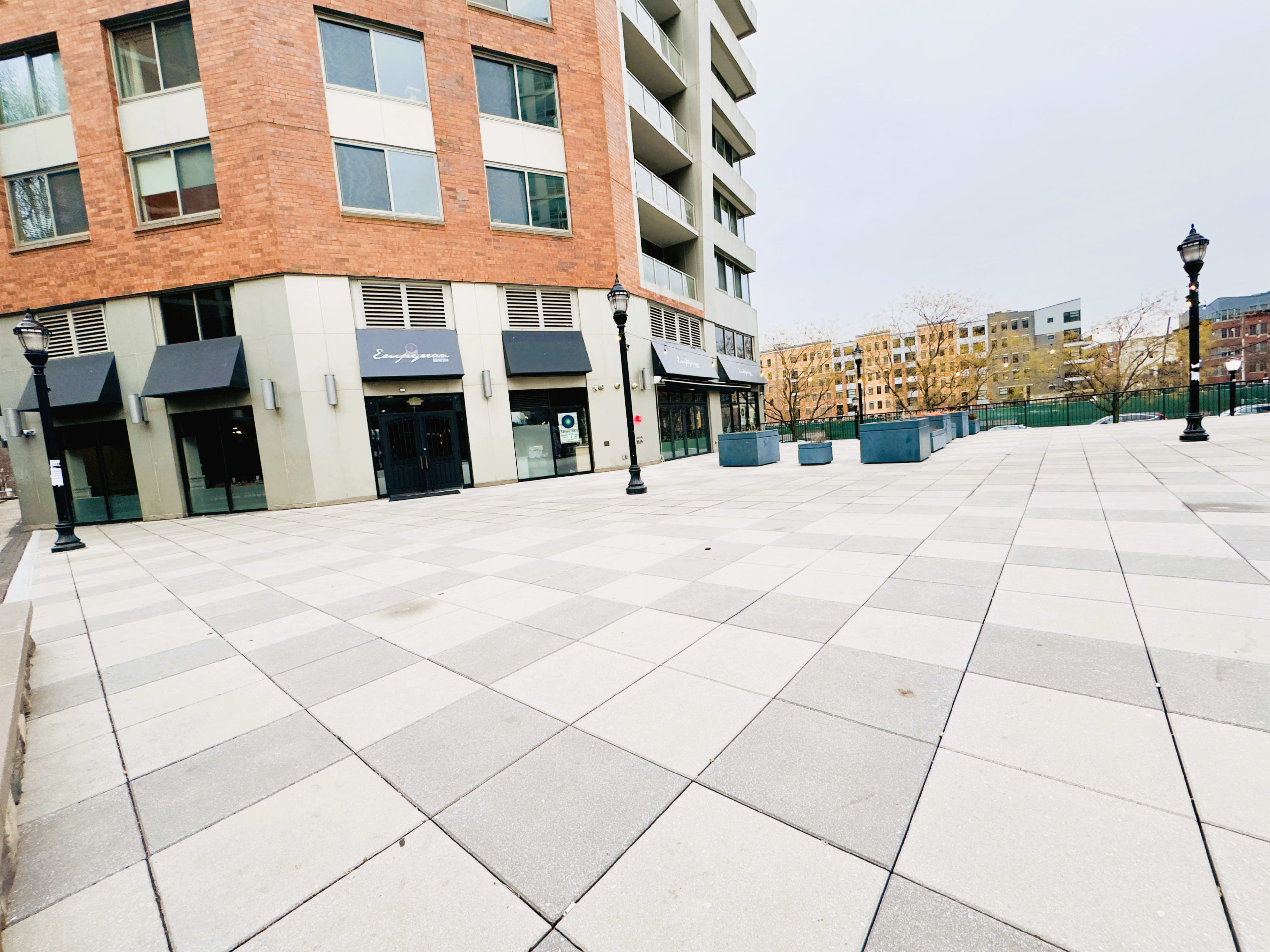The image depicts a spacious, paved plaza in front of a multi-story brick building, featuring lampposts and several planters.