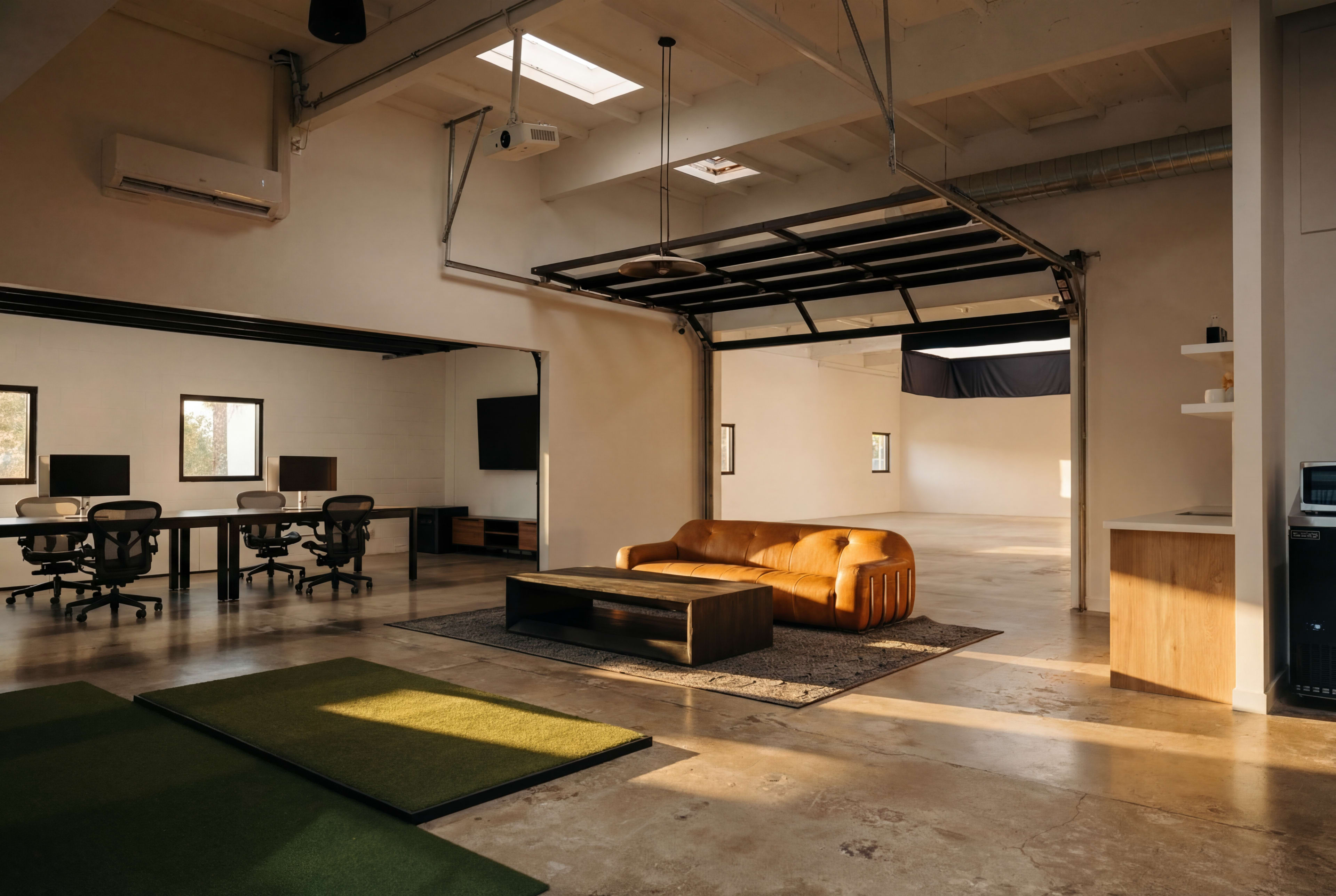 The image features a modern, open-concept workspace with a brown couch, a coffee table, and two desks equipped with computer monitors.