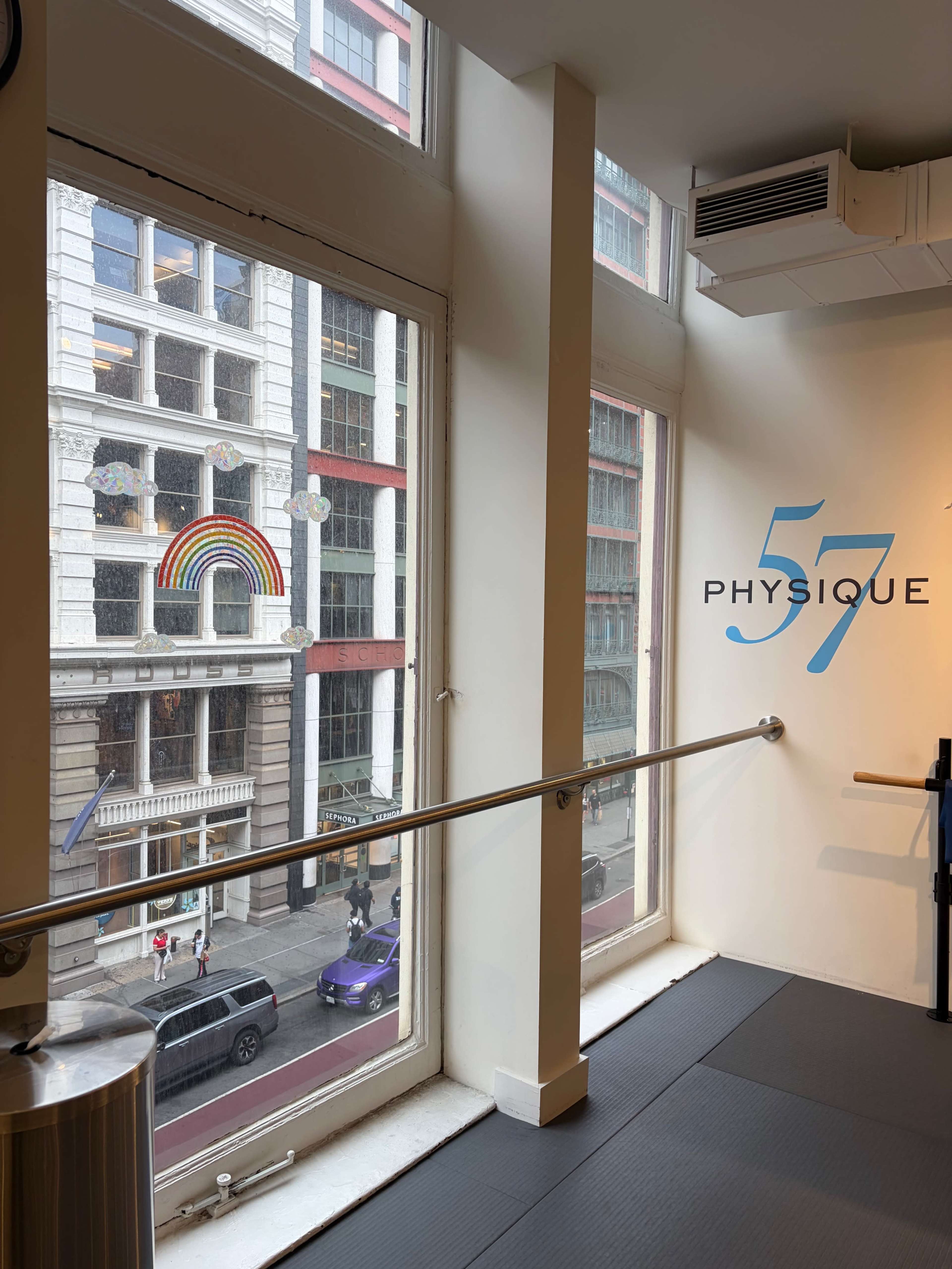 The image shows a fitness studio interior with large windows overlooking a street featuring a rainbow mural on a building across the way.